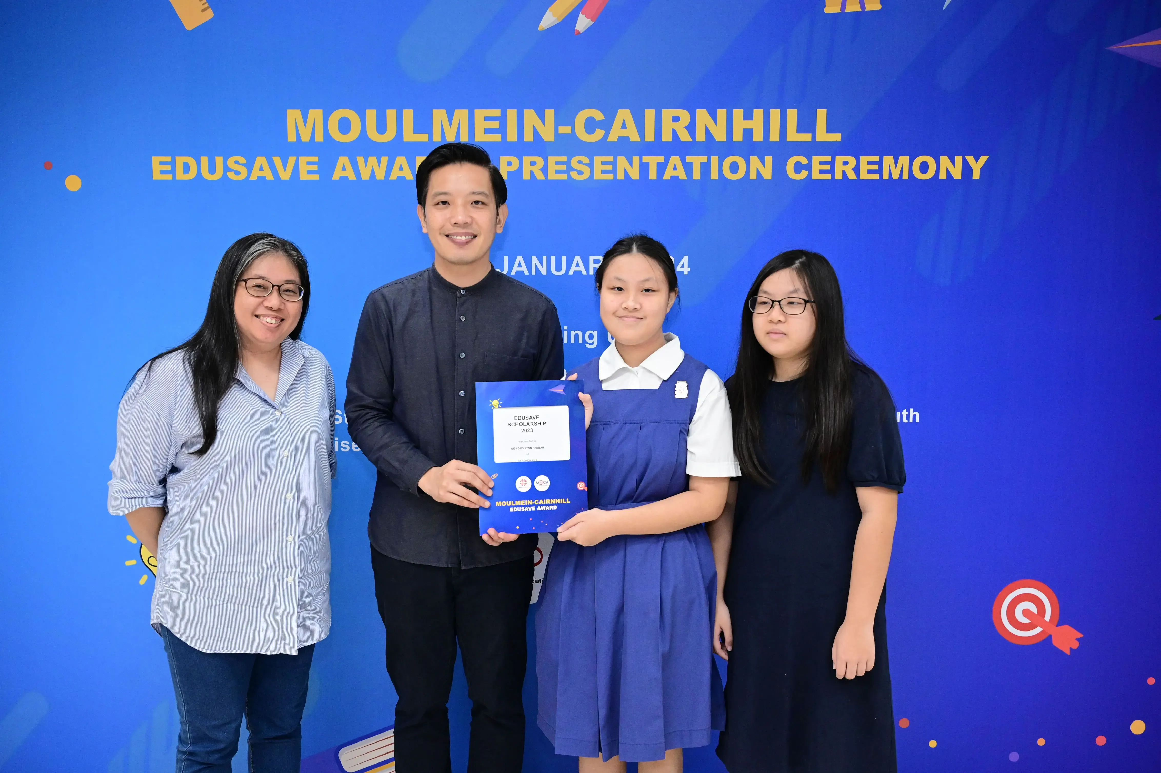 Four people pose at the Moulmein-Cairnhill Edusave Award Presentation Ceremony, with a student holding a scholarship certificate.