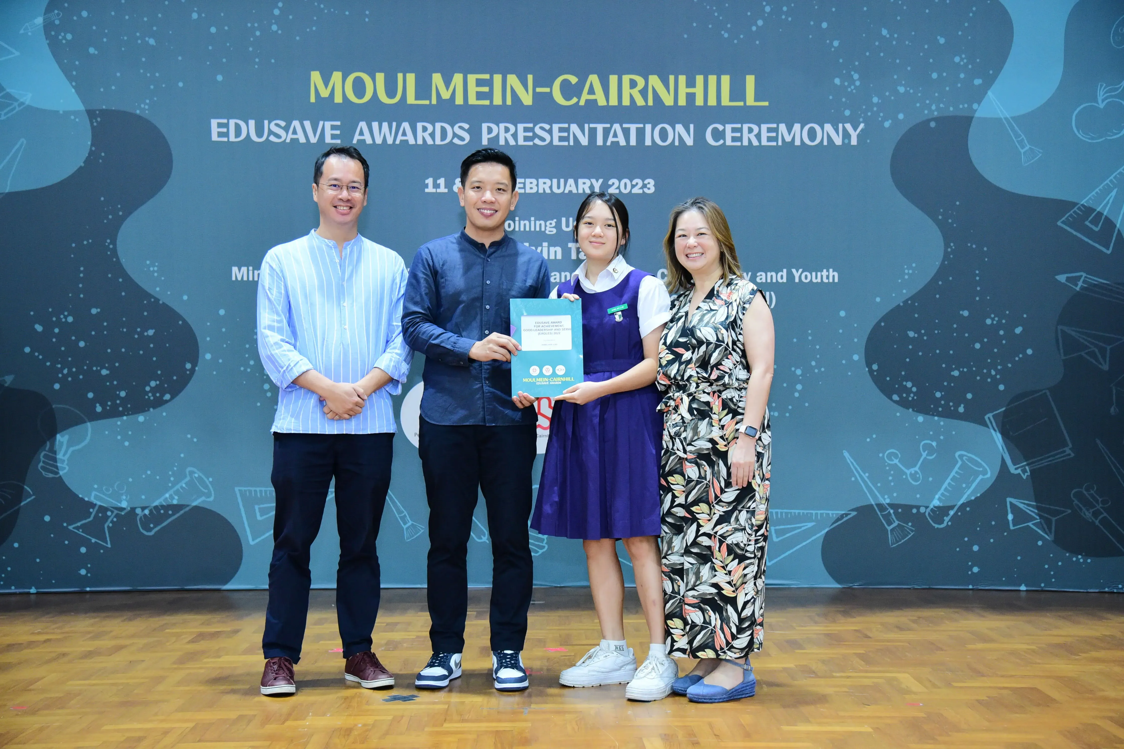 Four people pose at the Moulmein-Cairnhill Edusave Awards Presentation Ceremony, with a student holding a certificate.