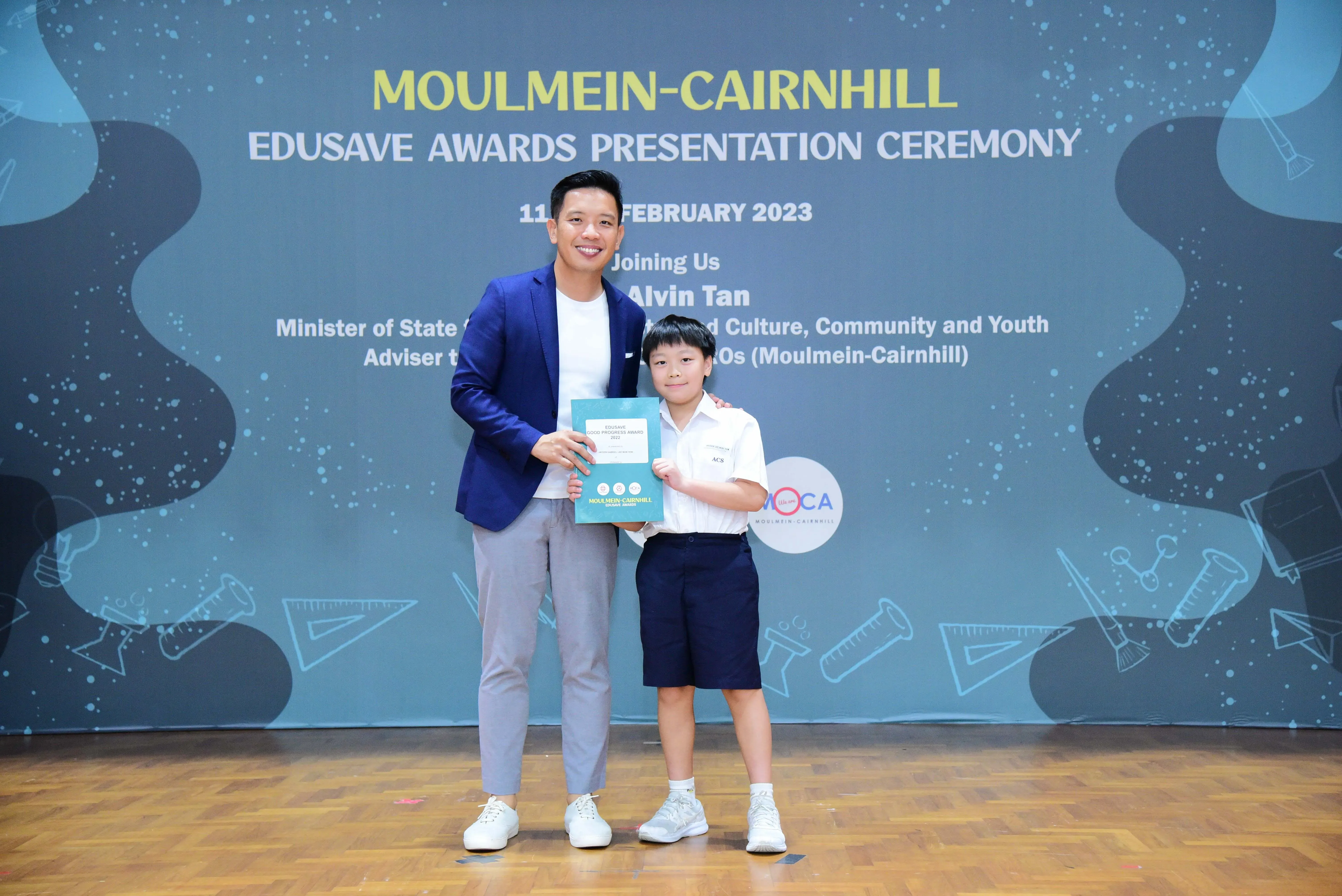 A man in a blue blazer and a young boy in school uniform hold a certificate at the Moulmein-Cairnhill Edusave Awards 2023.