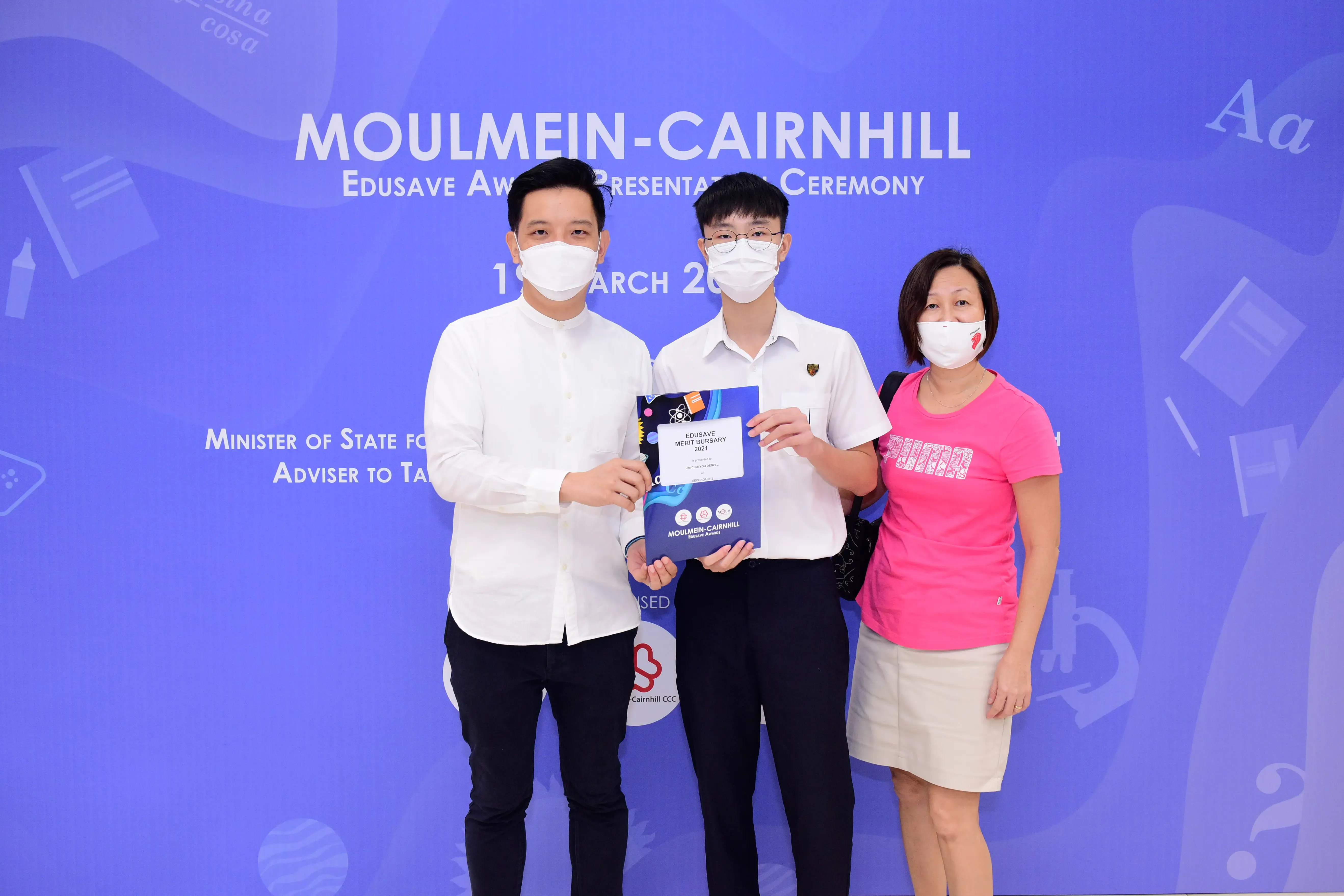 Three masked people pose at Moulmein-Cairnhill Edusave Award Ceremony, with a student holding a Merit Bursary folder.