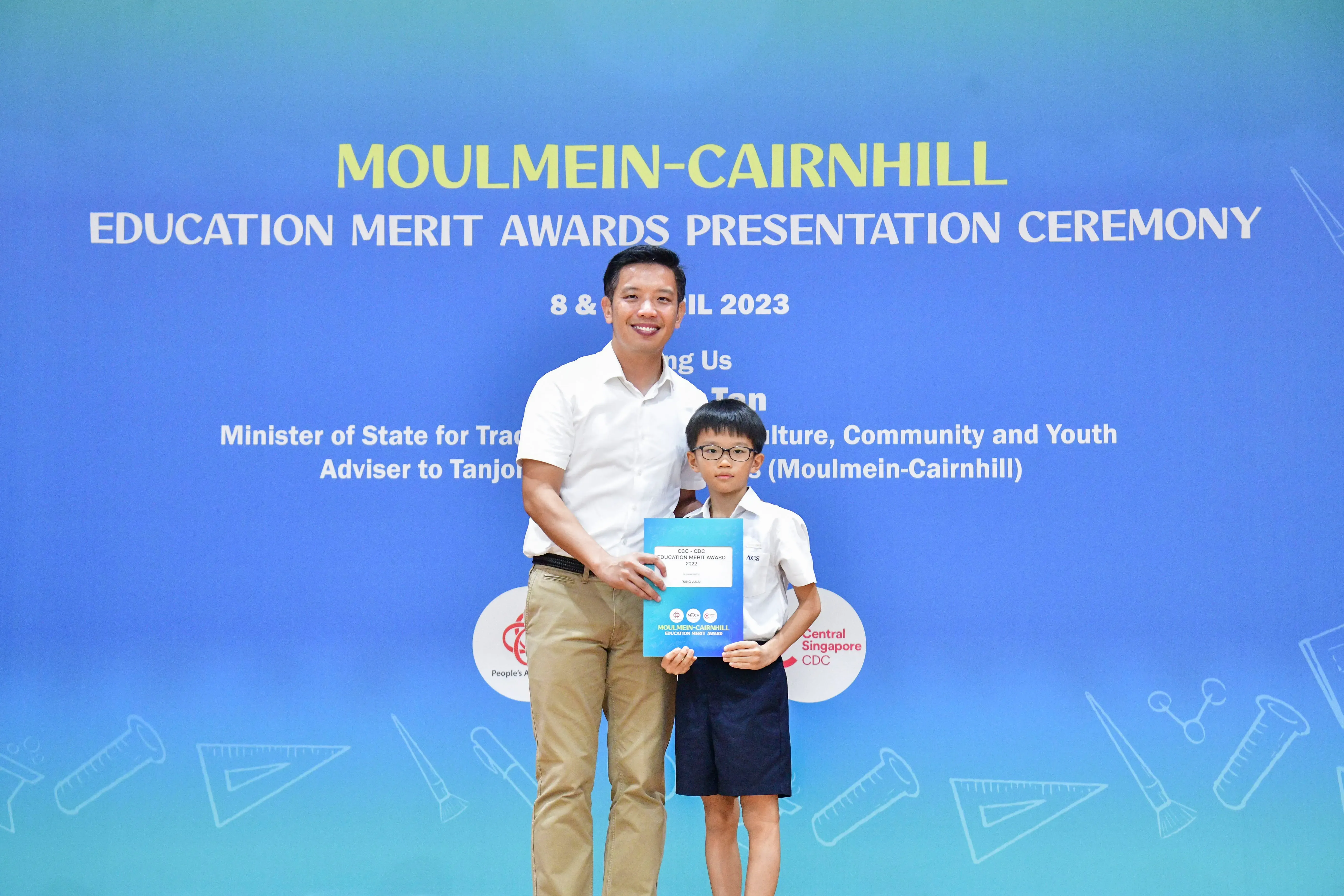 An adult and a young boy hold an award certificate in front of a Moulmein-Cairnhill Education Merit Awards 2023 banner.