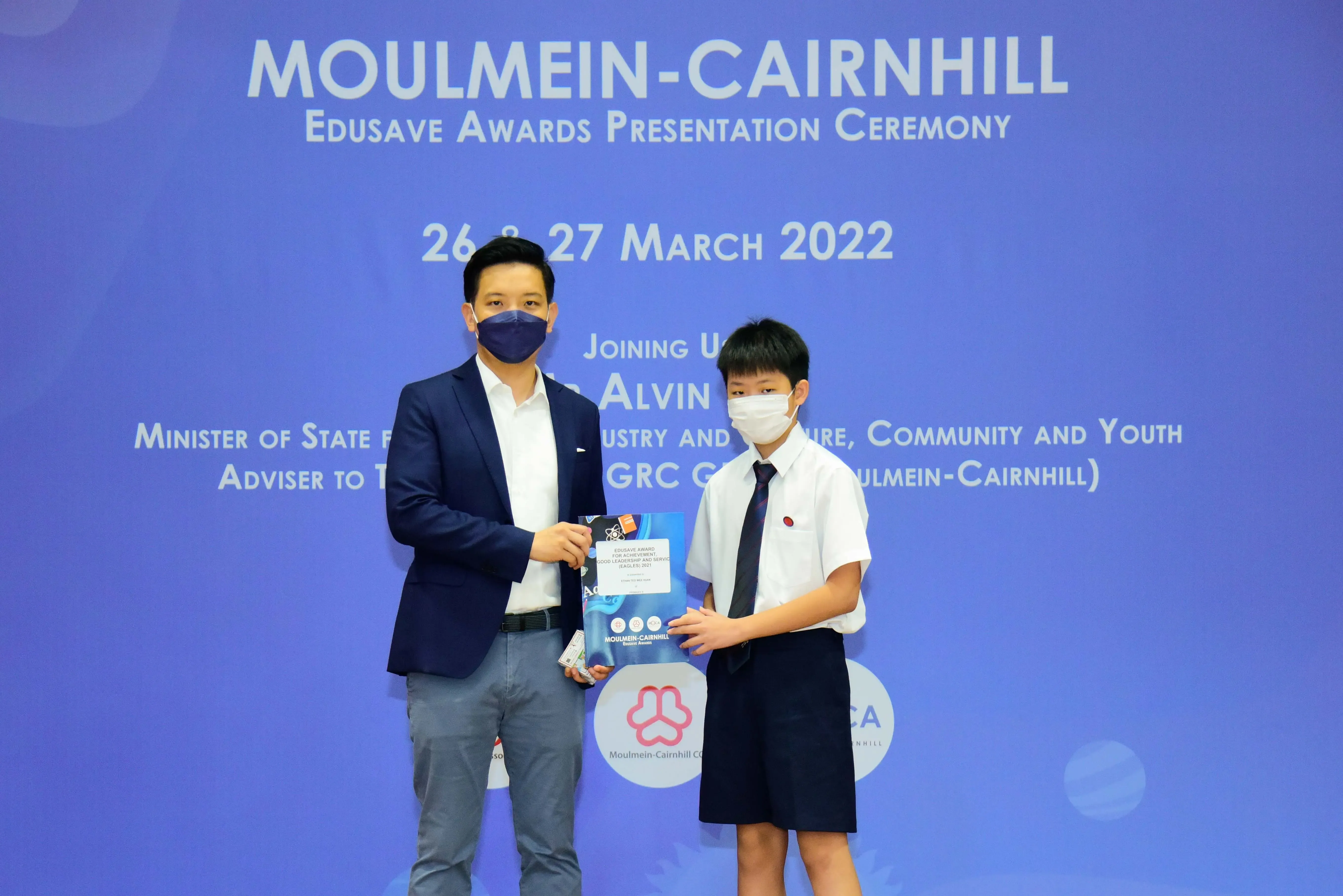 An adult in a suit and a young student in uniform hold an award at the Moulmein-Cairnhill Edusave ceremony, March 2022.