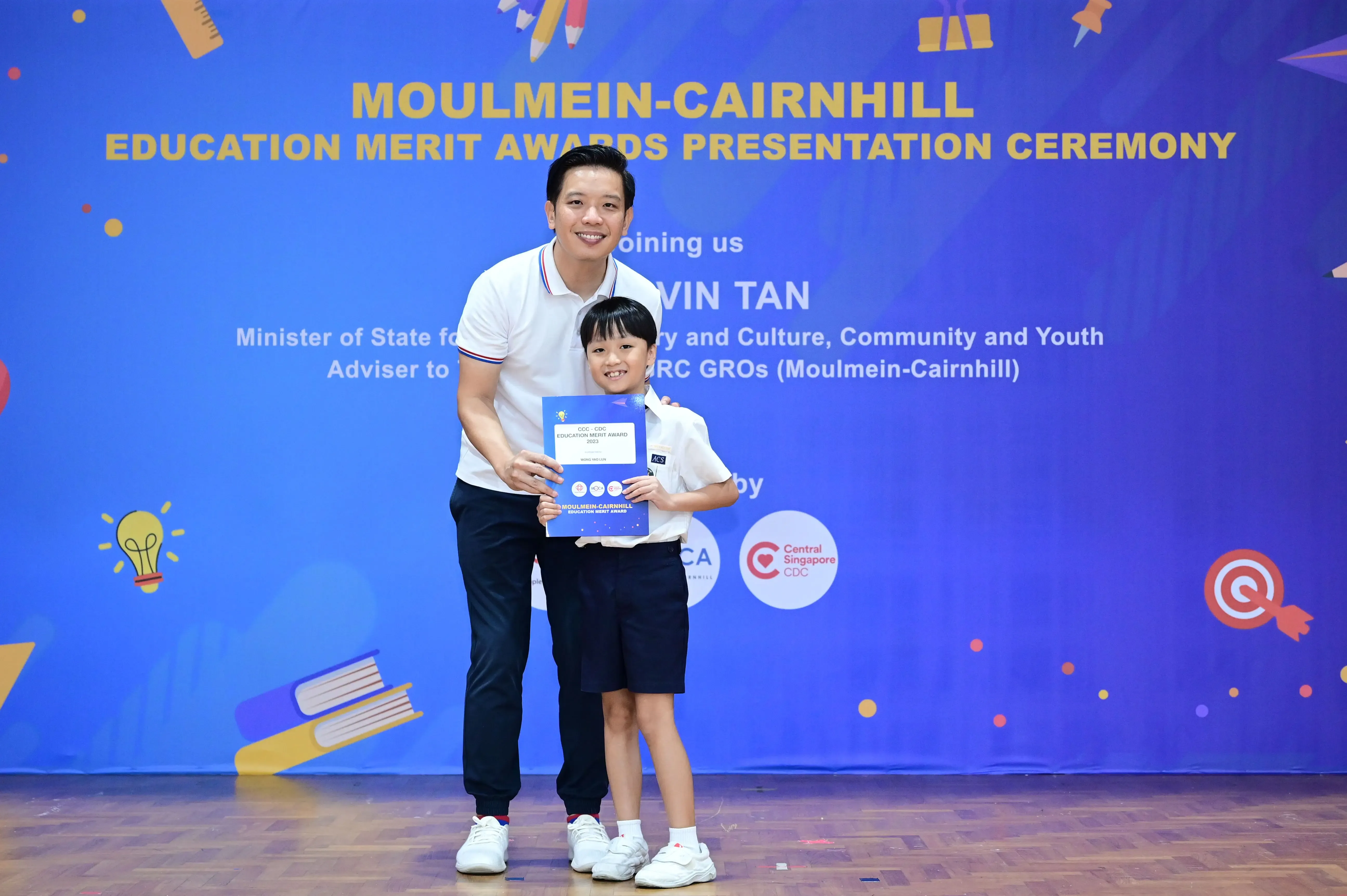 Adult man and young child pose together holding an award certificate at the Moulmein-Cairnhill Education Merit Awards ceremony.