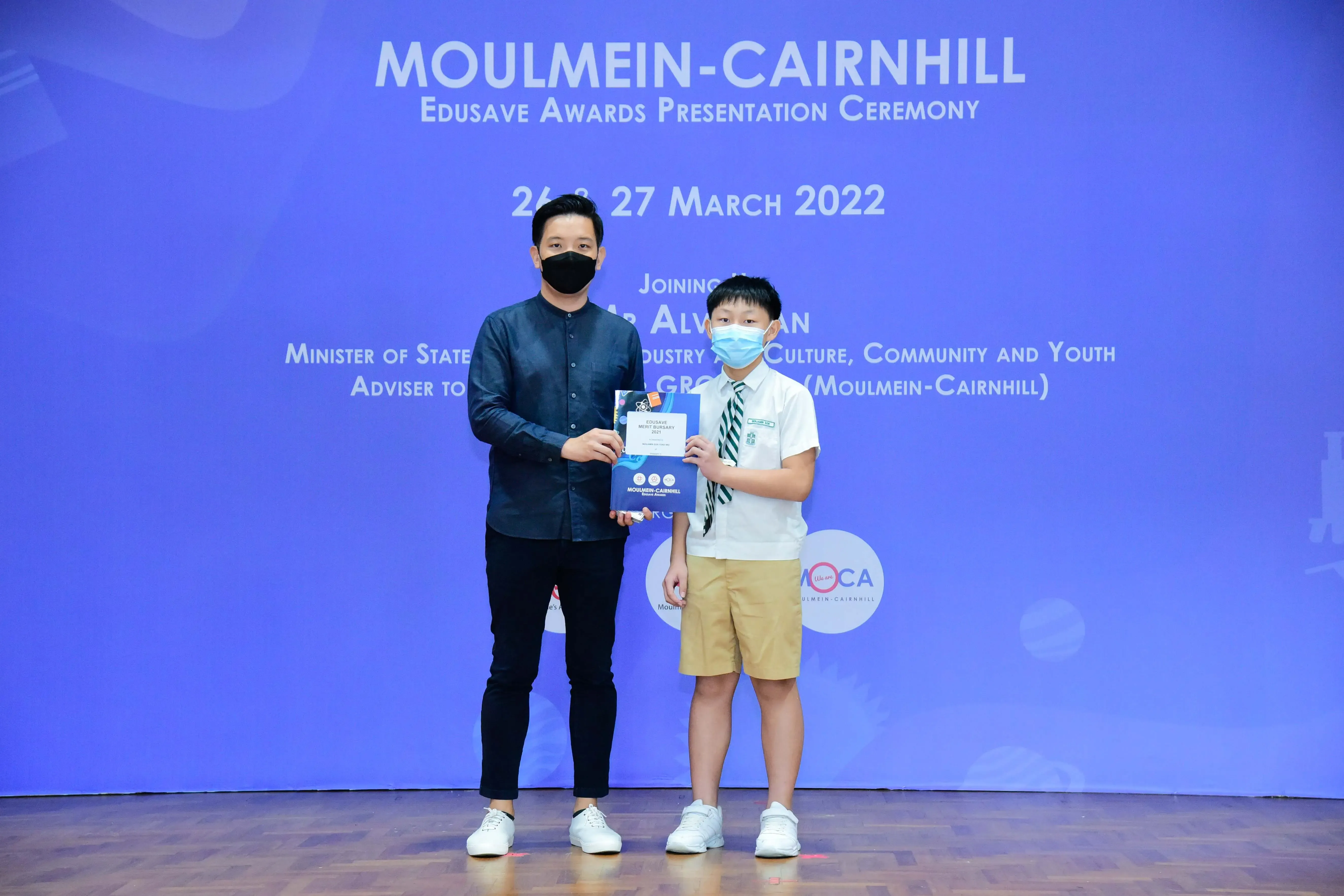An adult and a masked student hold an award certificate at the Moulmein-Cairnhill Edusave Awards Ceremony, March 2022.