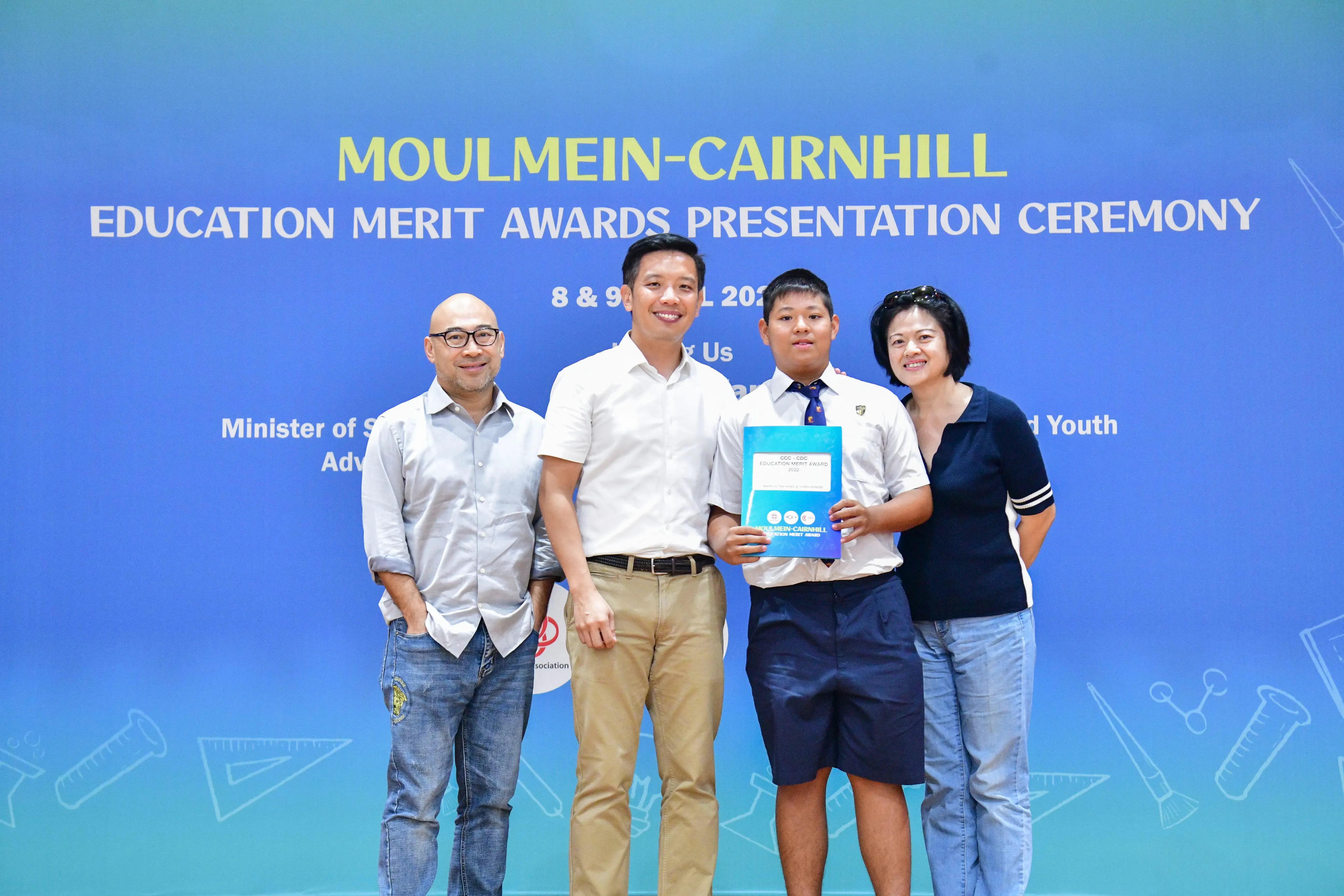 Four people pose before a blue Moulmein-Cairnhill Education Merit Awards banner; a boy holds a certificate.