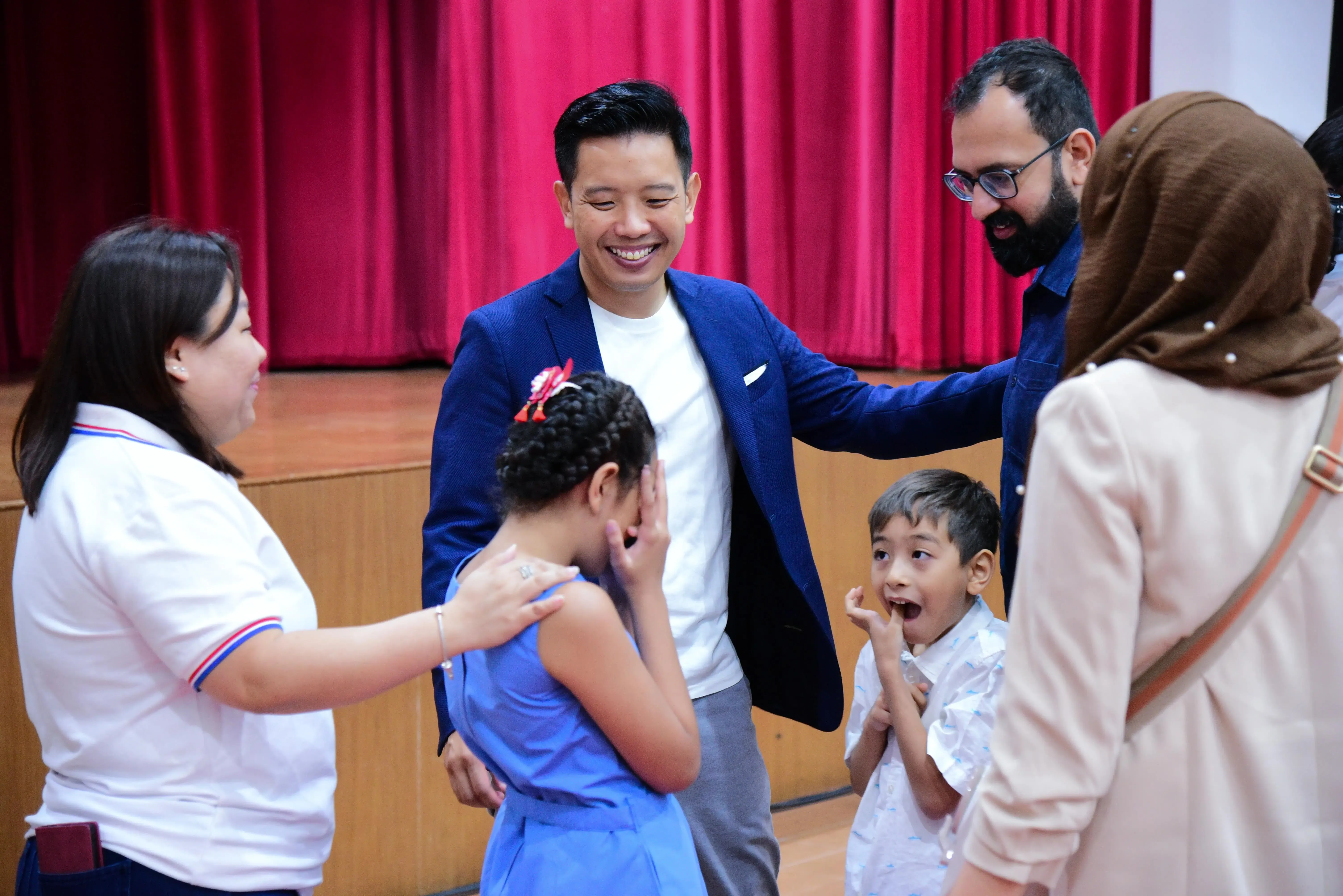 A smiling man in a blue blazer interacts with two children and three adults near a red curtain stage.