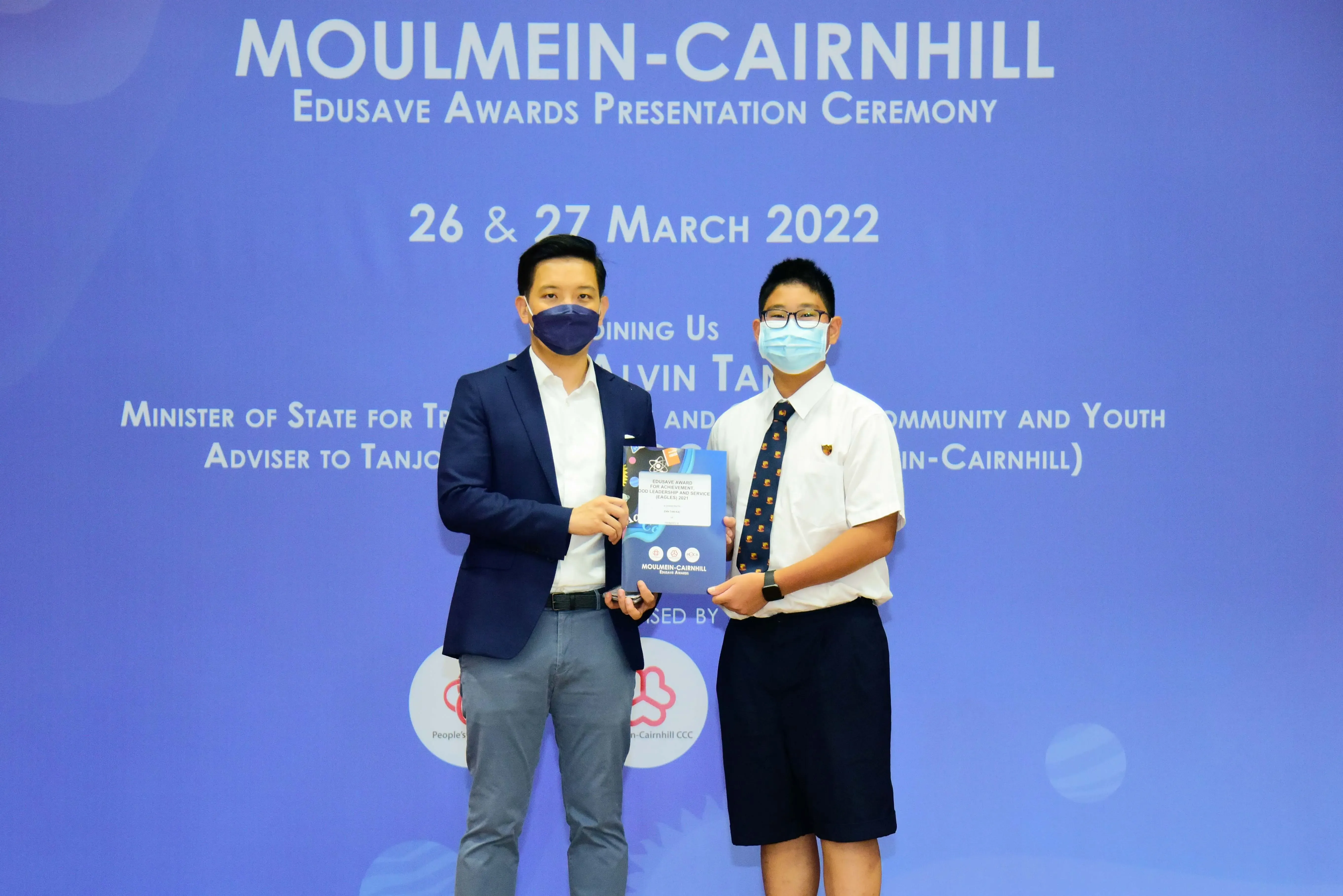 Two masked individuals hold an award folder at the Moulmein-Cairnhill Edusave Awards Presentation Ceremony, March 2022.