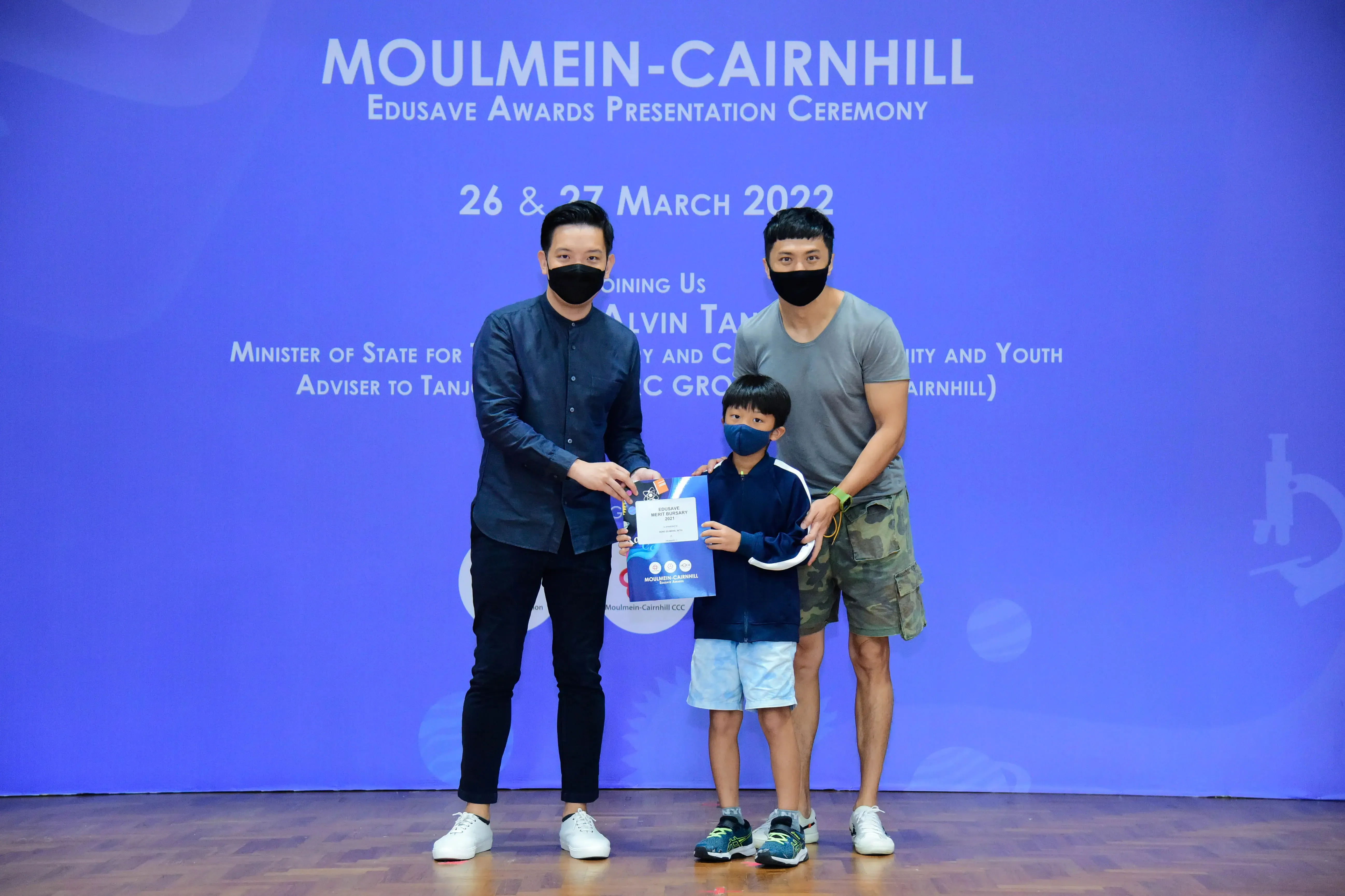 Two masked adults and a masked child hold an award at the Moulmein-Cairnhill Edusave Awards Ceremony, March 2022.