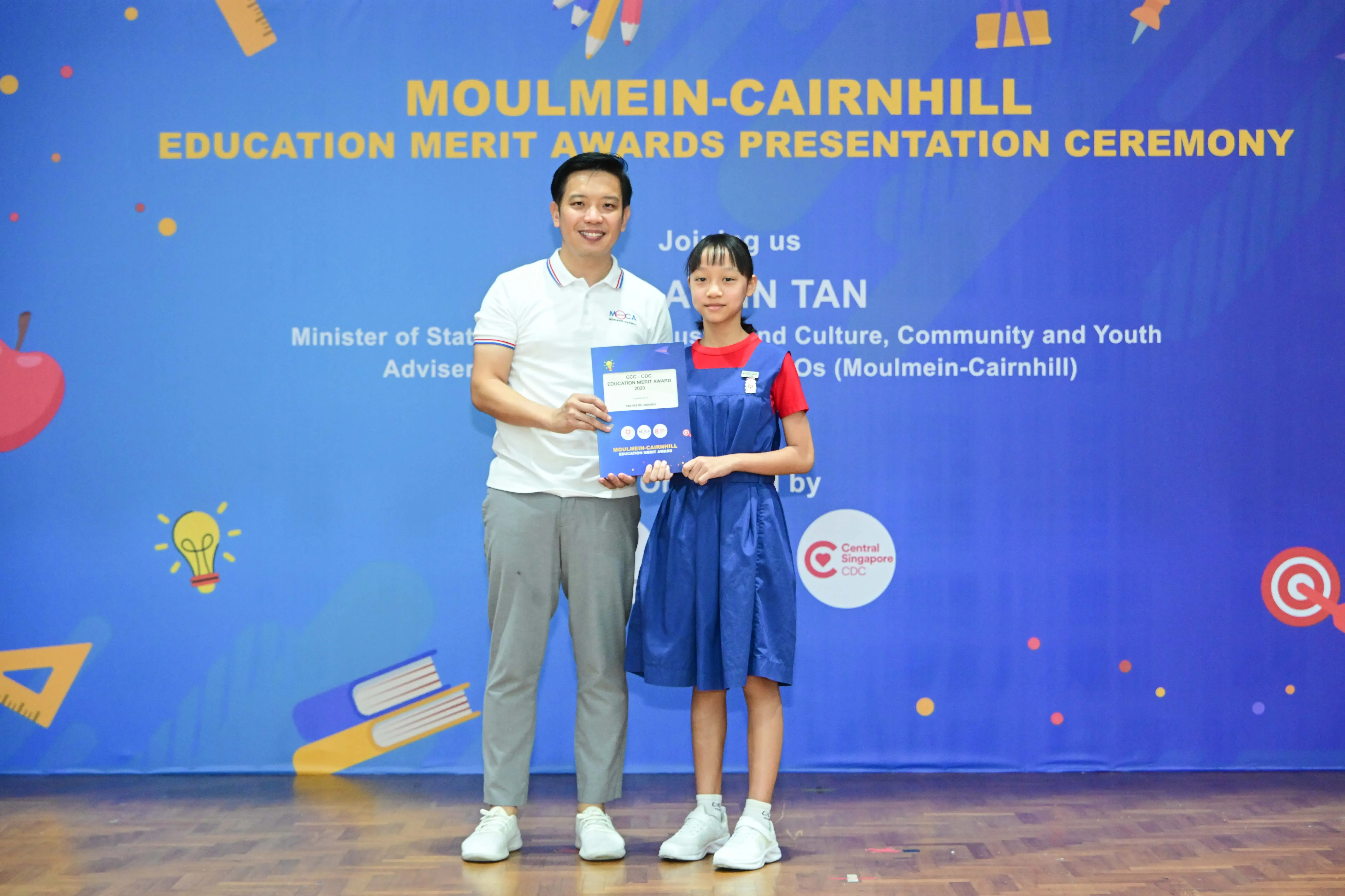 A man and a young girl hold a certificate together in front of a Moulmein-Cairnhill Education Merit Awards banner.
