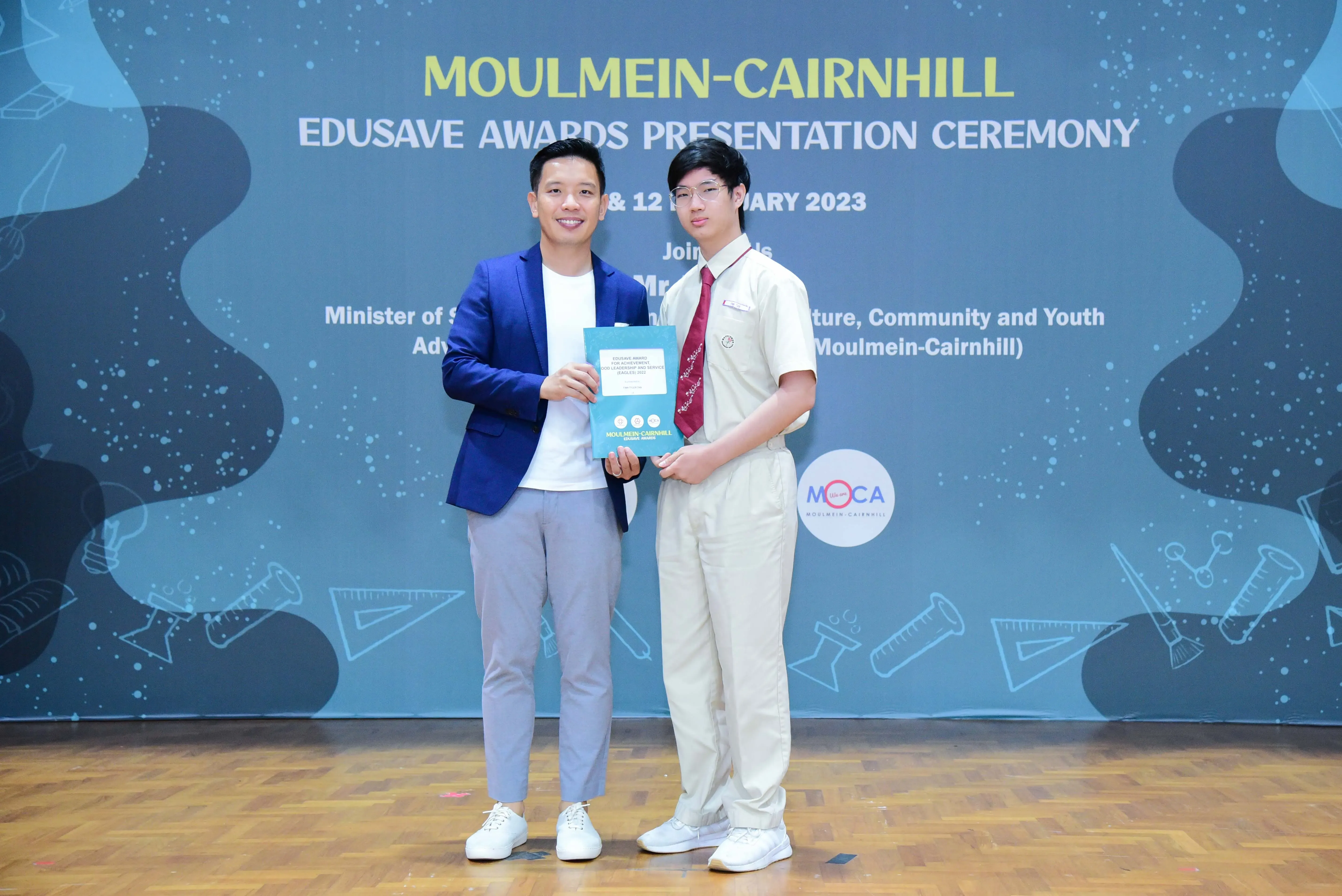 Two men hold a certificate together on stage at the Moulmein-Cairnhill Edusave Awards Presentation Ceremony 2023.