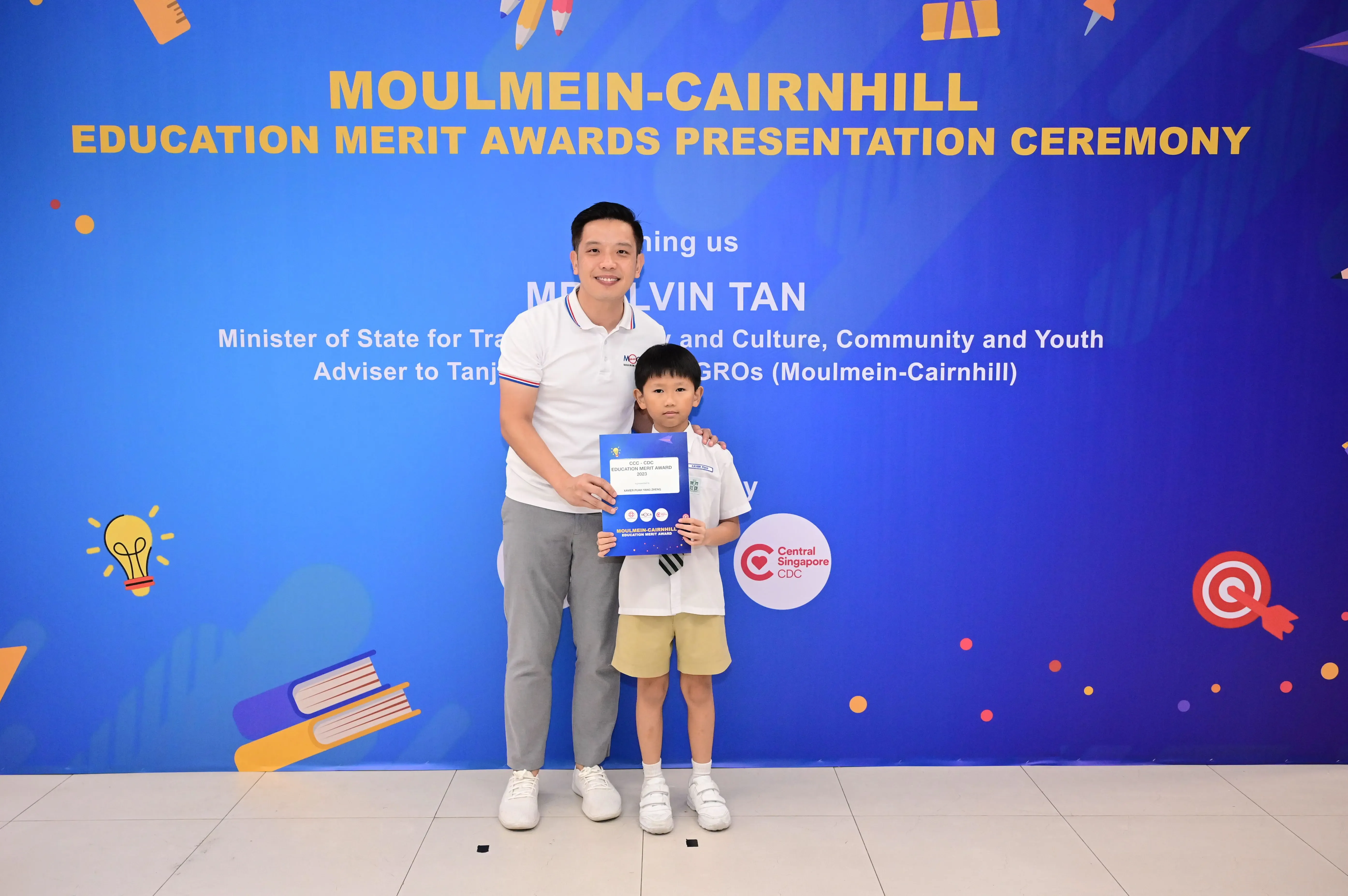 A man and a young boy hold an award certificate in front of a Moulmein-Cairnhill Education Merit Awards banner.