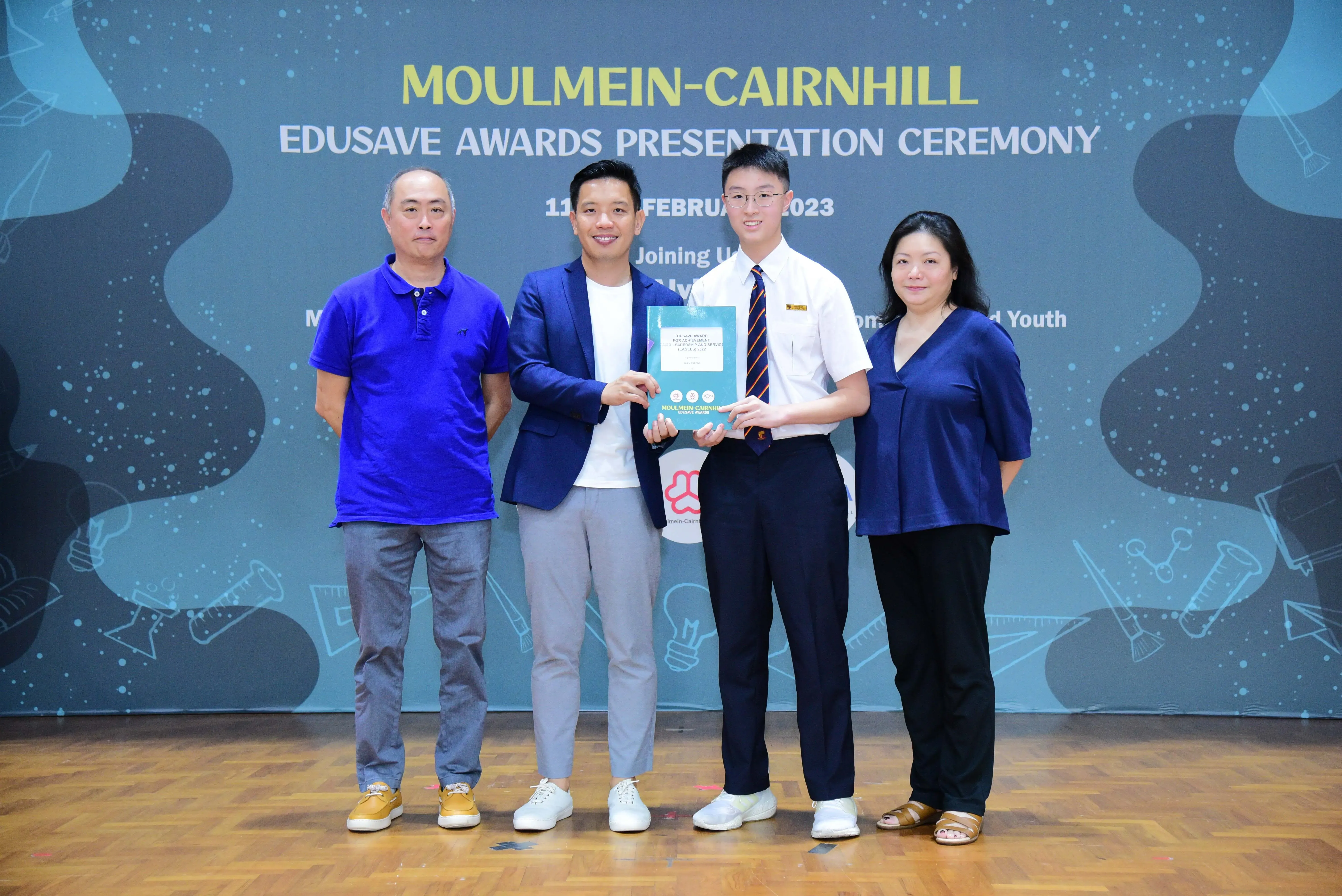 Four people pose at the Moulmein-Cairnhill Edusave Awards Presentation Ceremony, with two holding a teal award booklet.