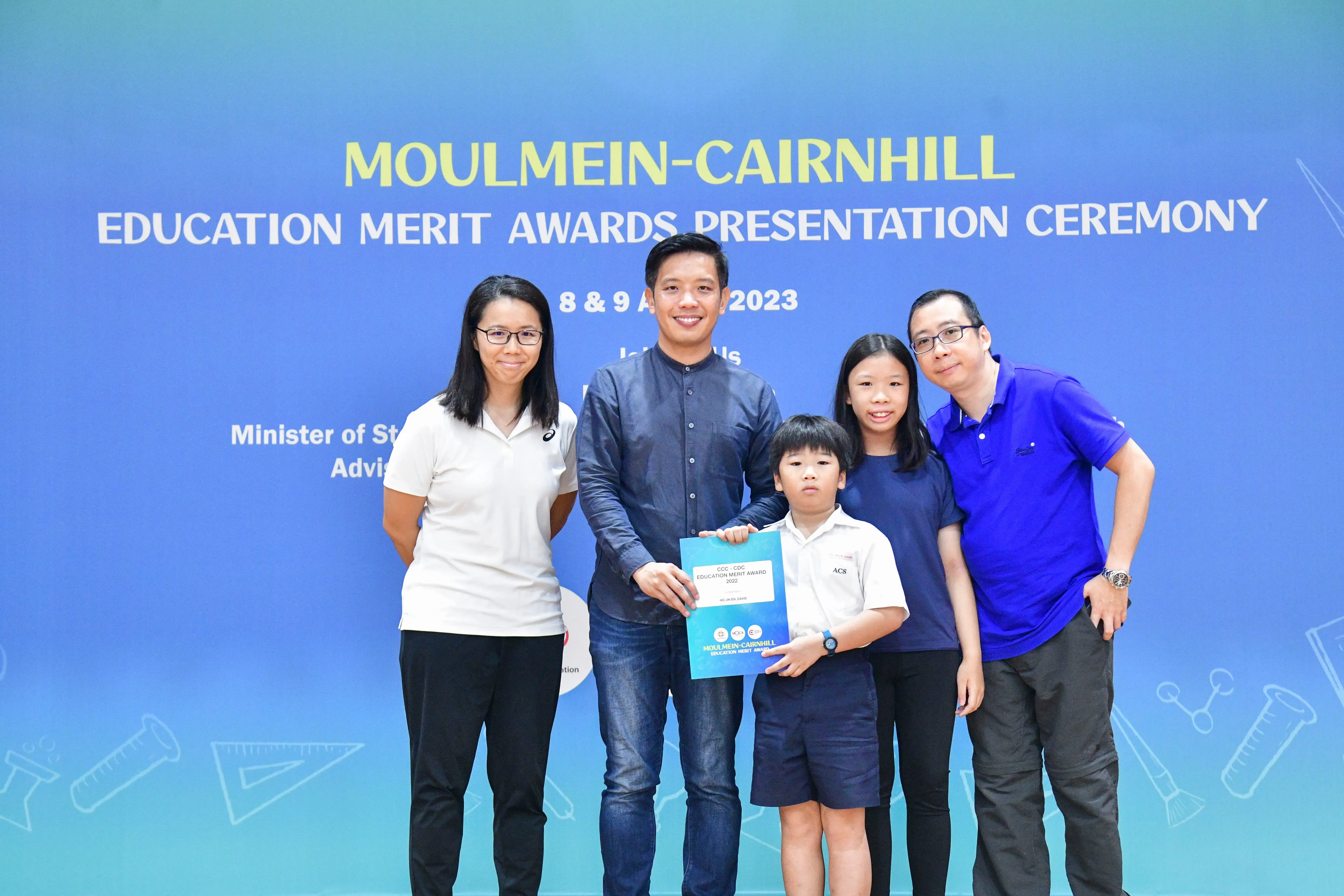 Five people pose before a blue Moulmein-Cairnhill Education Merit Awards Presentation Ceremony backdrop, child holding award.
