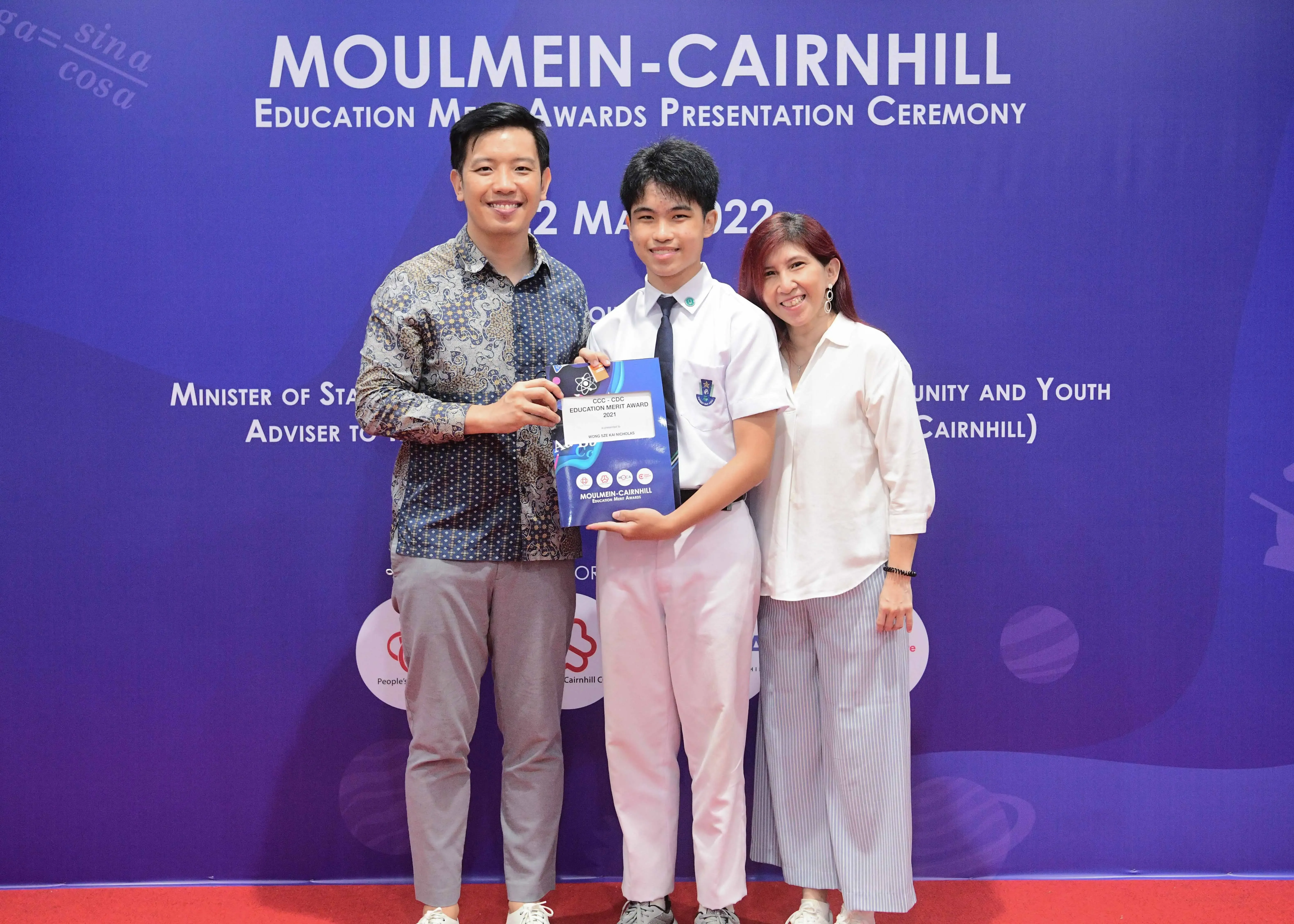 Three people smile holding an award at the Moulmein-Cairnhill Education Merit Awards Presentation Ceremony 2022.