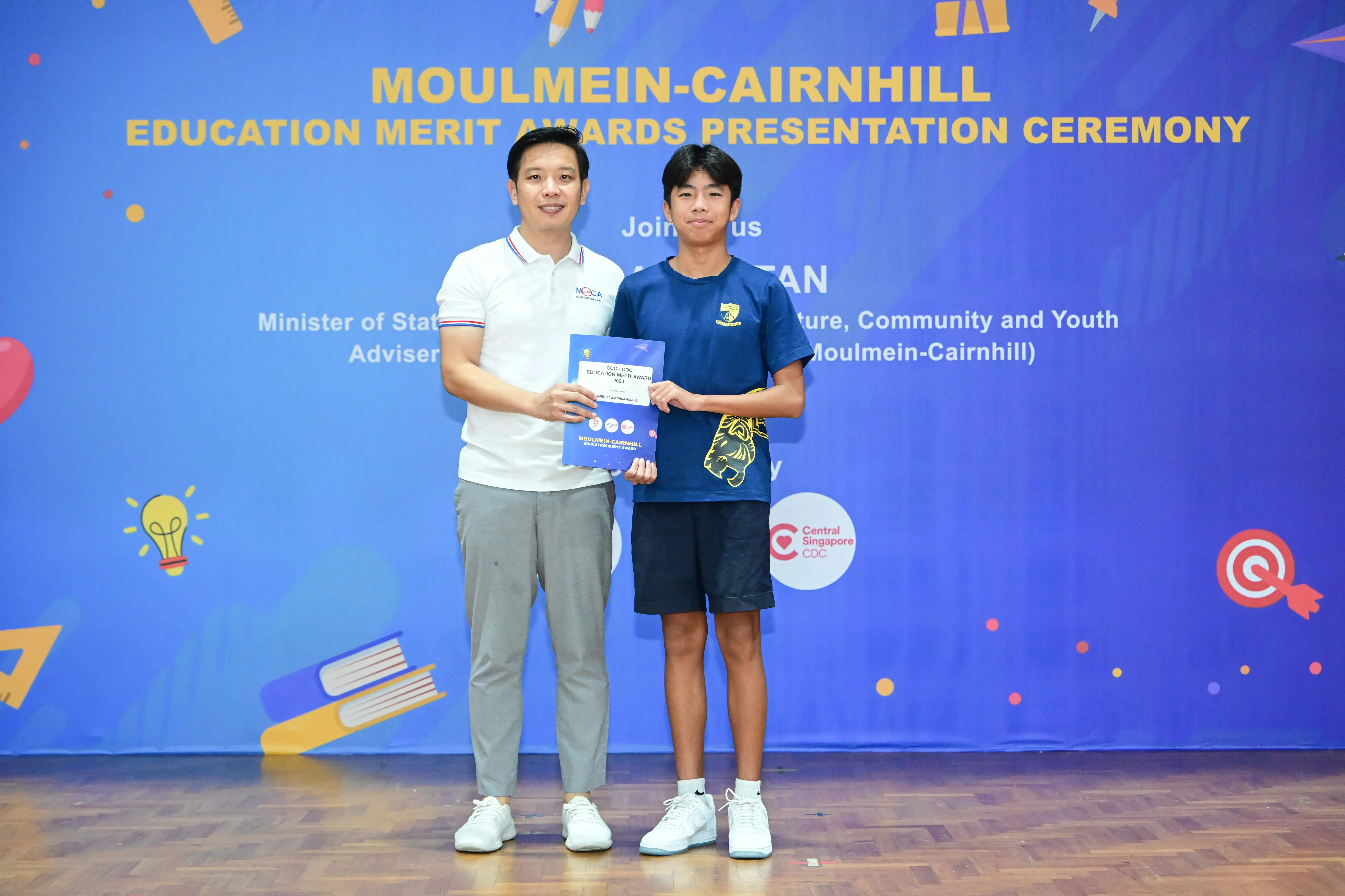 Two men pose holding a blue booklet at the Moulmein-Cairnhill Education Merit Awards Presentation Ceremony.