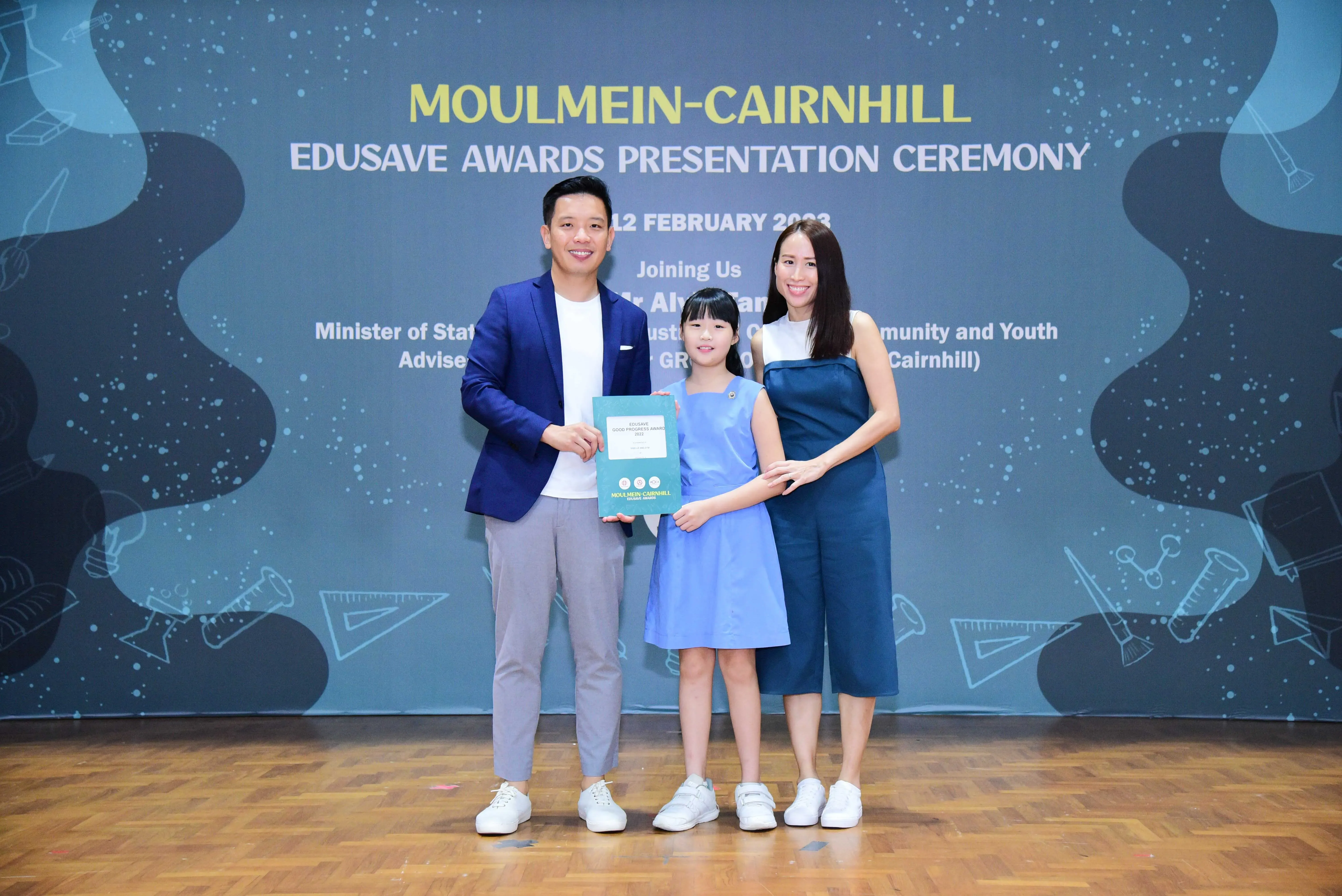 A man, young girl holding an award certificate, and woman pose at the Moulmein-Cairnhill Edusave Awards Ceremony backdrop.