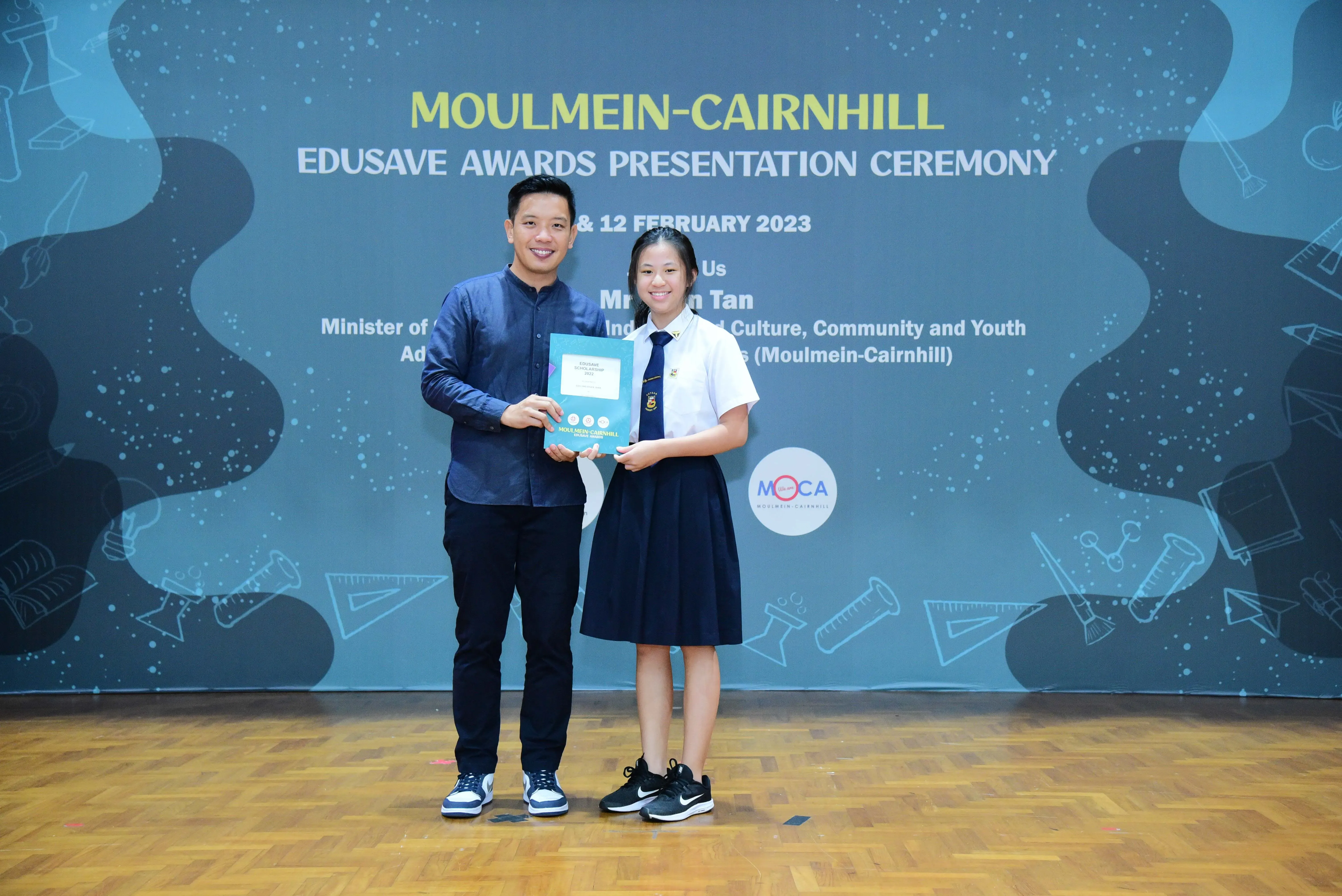 A man and a female student hold a certificate together at the Moulmein-Cairnhill Edusave Awards Ceremony 2023.
