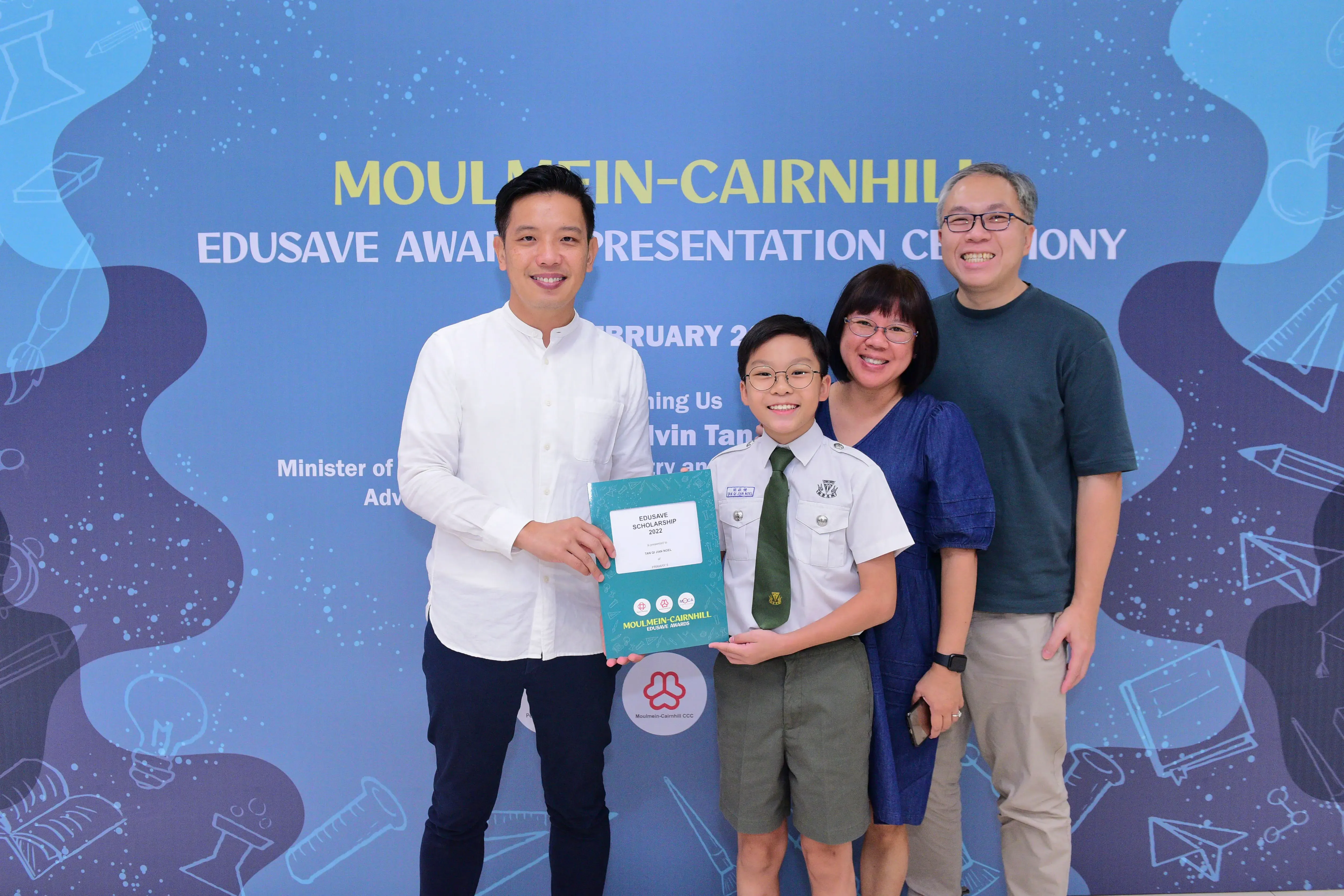 A boy in school uniform holds an Edusave Scholarship certificate with three smiling adults at an awards ceremony.