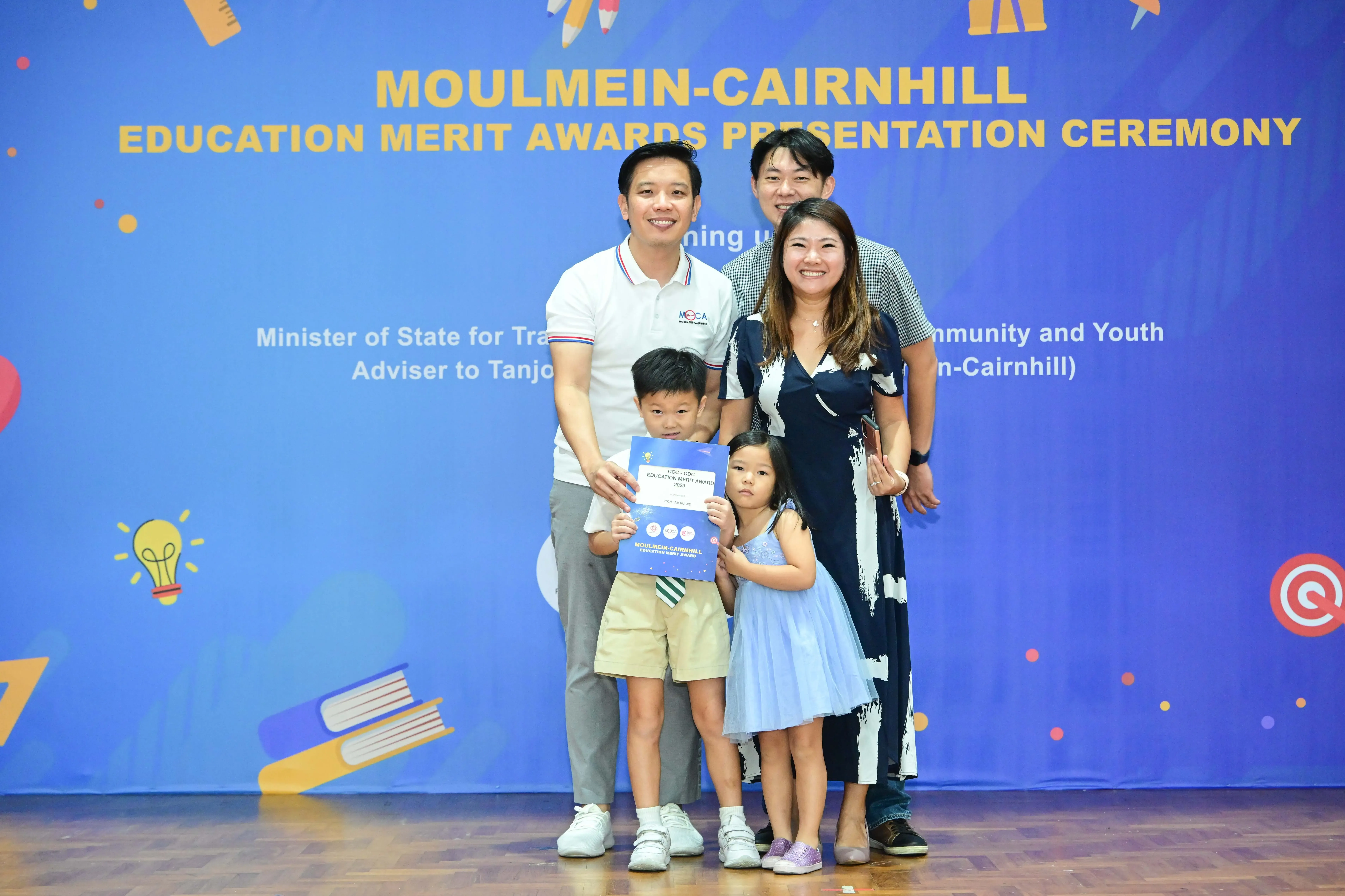Four adults and two young children pose with a certificate in front of a Moulmein-Cairnhill Education Merit Awards banner.