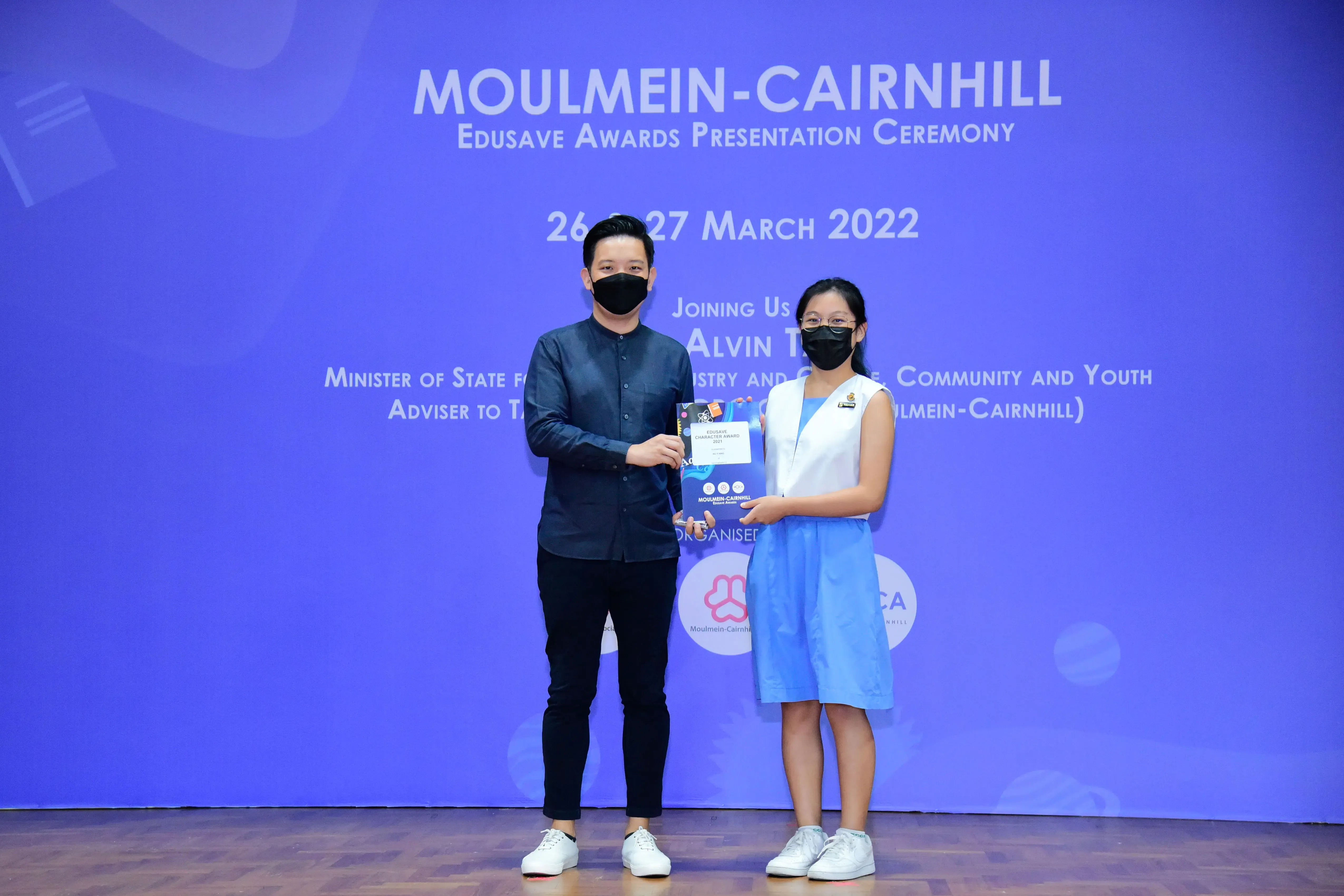Two masked individuals hold an award booklet on stage at the Moulmein-Cairnhill Edusave Awards Ceremony, March 2022.