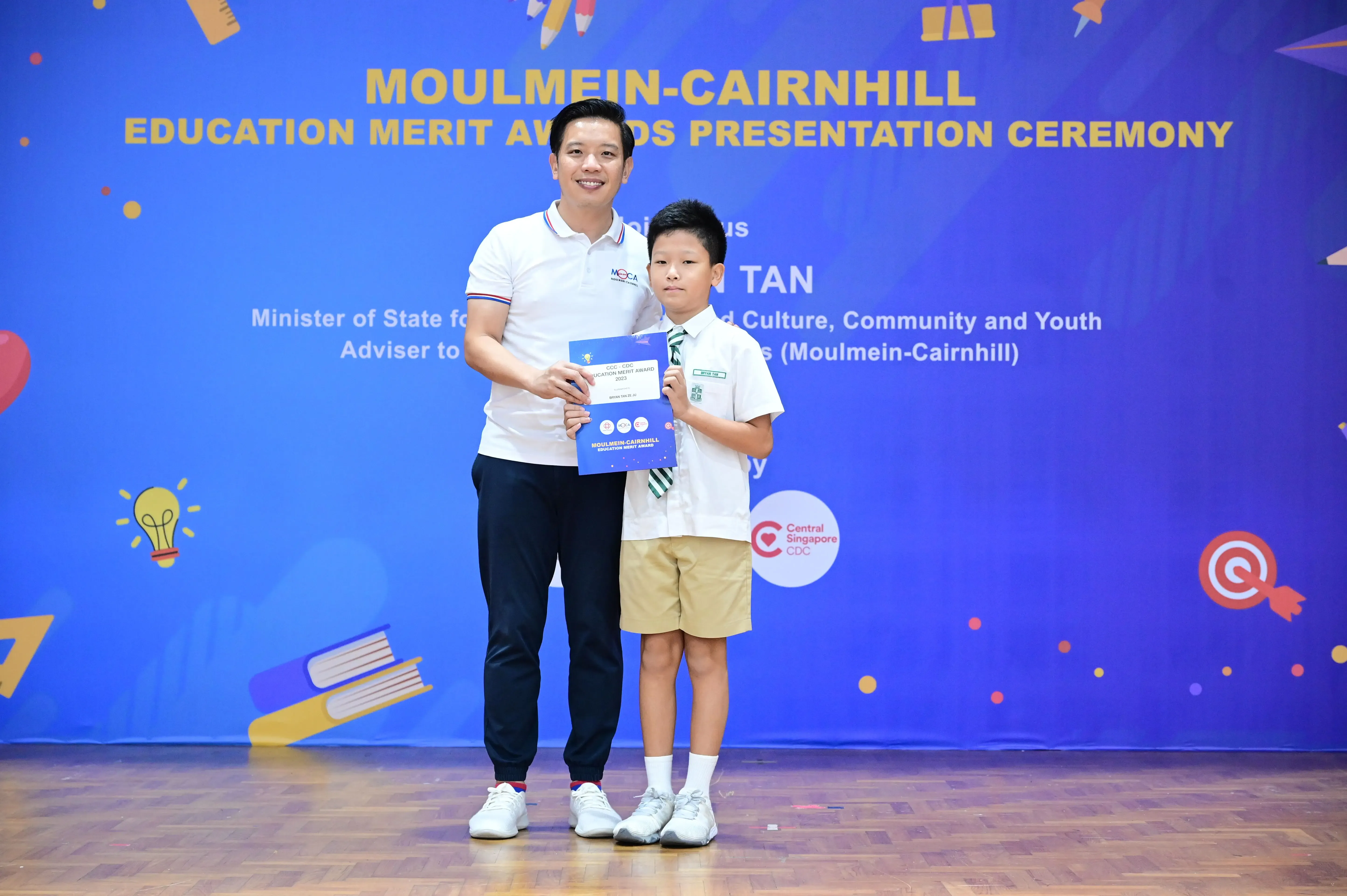 A man and a young boy hold a certificate together at the Moulmein-Cairnhill Education Merit Awards ceremony.