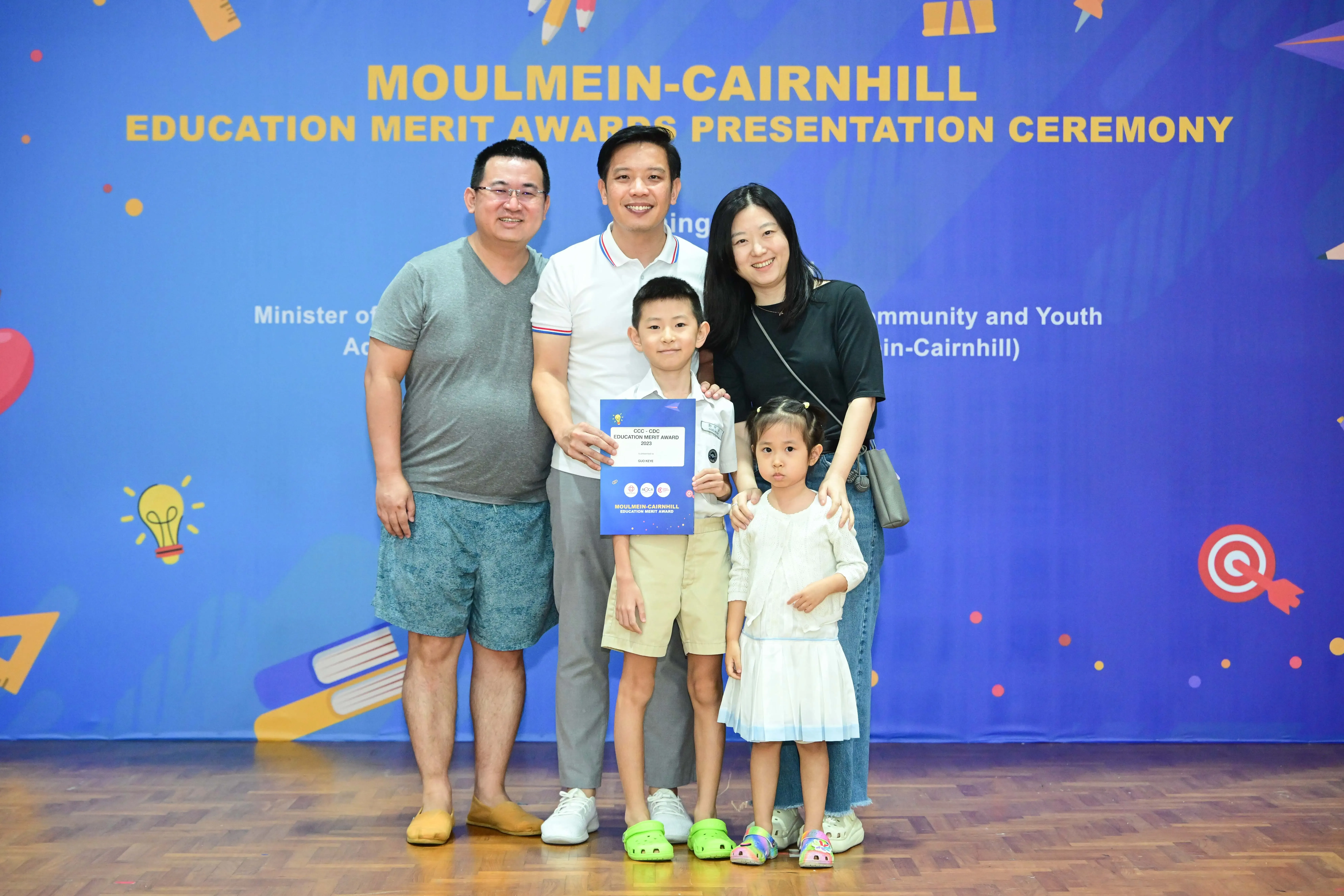 Family of five posing with an award certificate in front of a Moulmein-Cairnhill Education Merit Awards banner.