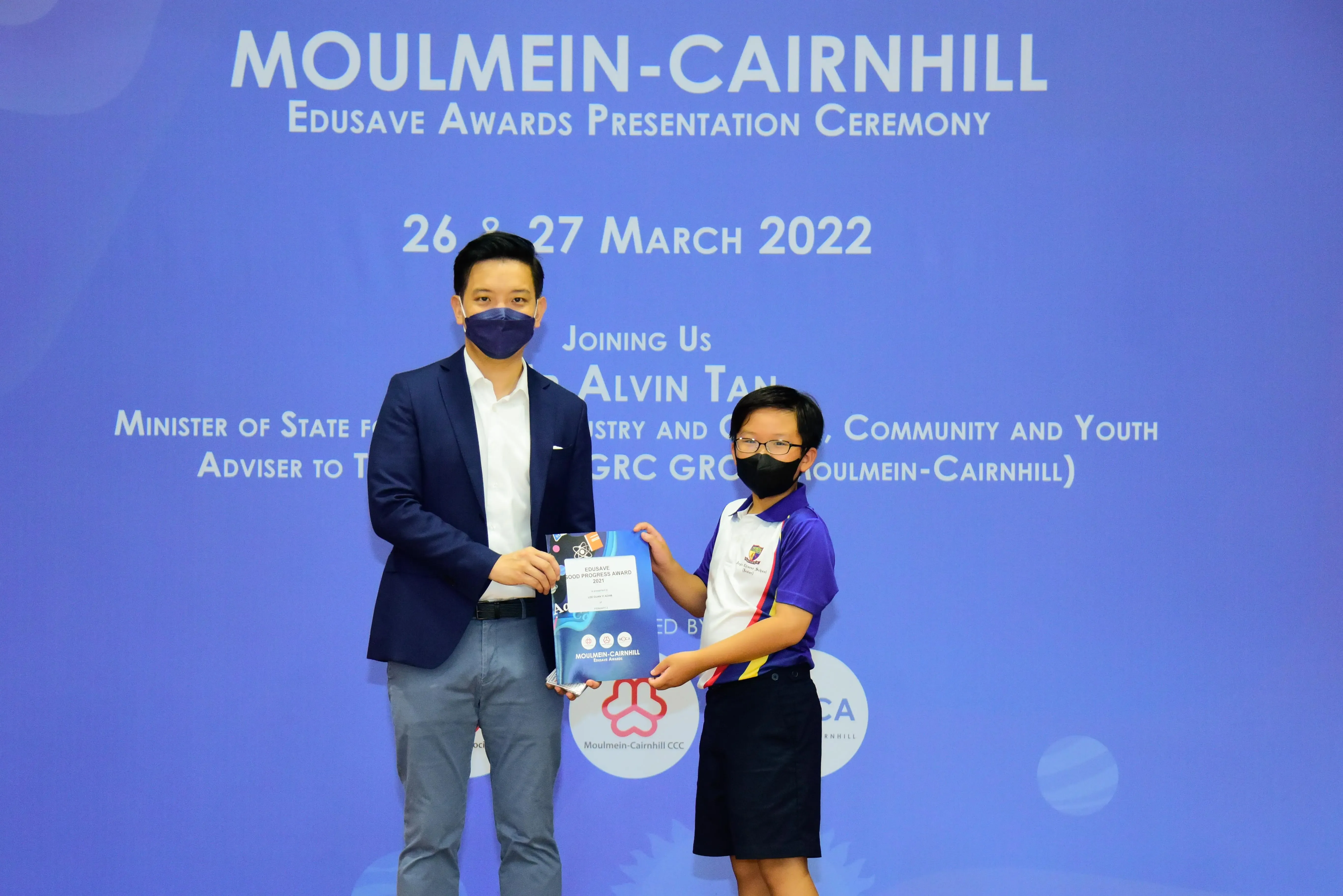 An adult and a child in masks hold an award certificate at the Moulmein-Cairnhill Edusave Awards Ceremony, March 2022.