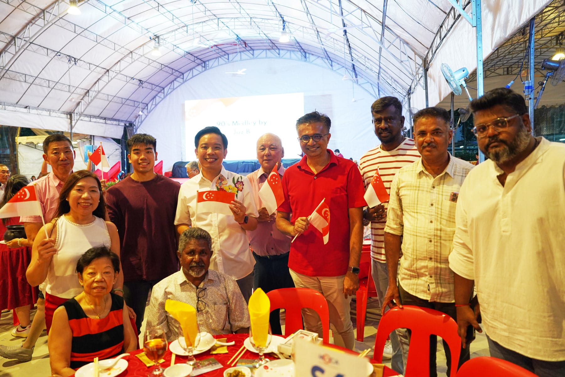 Group of people smiling and holding small Singapore flags at a festive event inside a white tent.