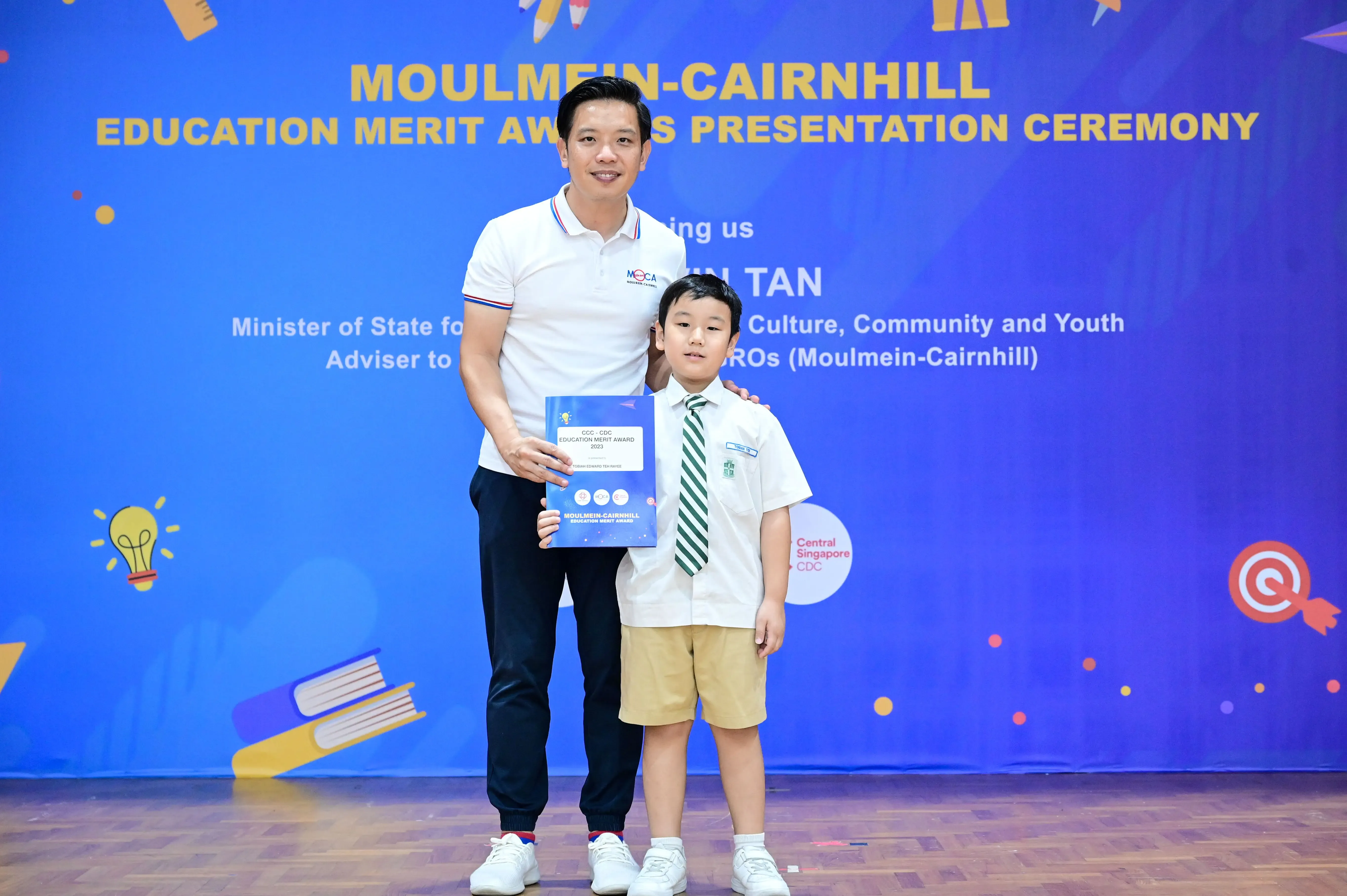 A man and a young boy in school uniform hold an award certificate at the Moulmein-Cairnhill Education Merit Awards ceremony.