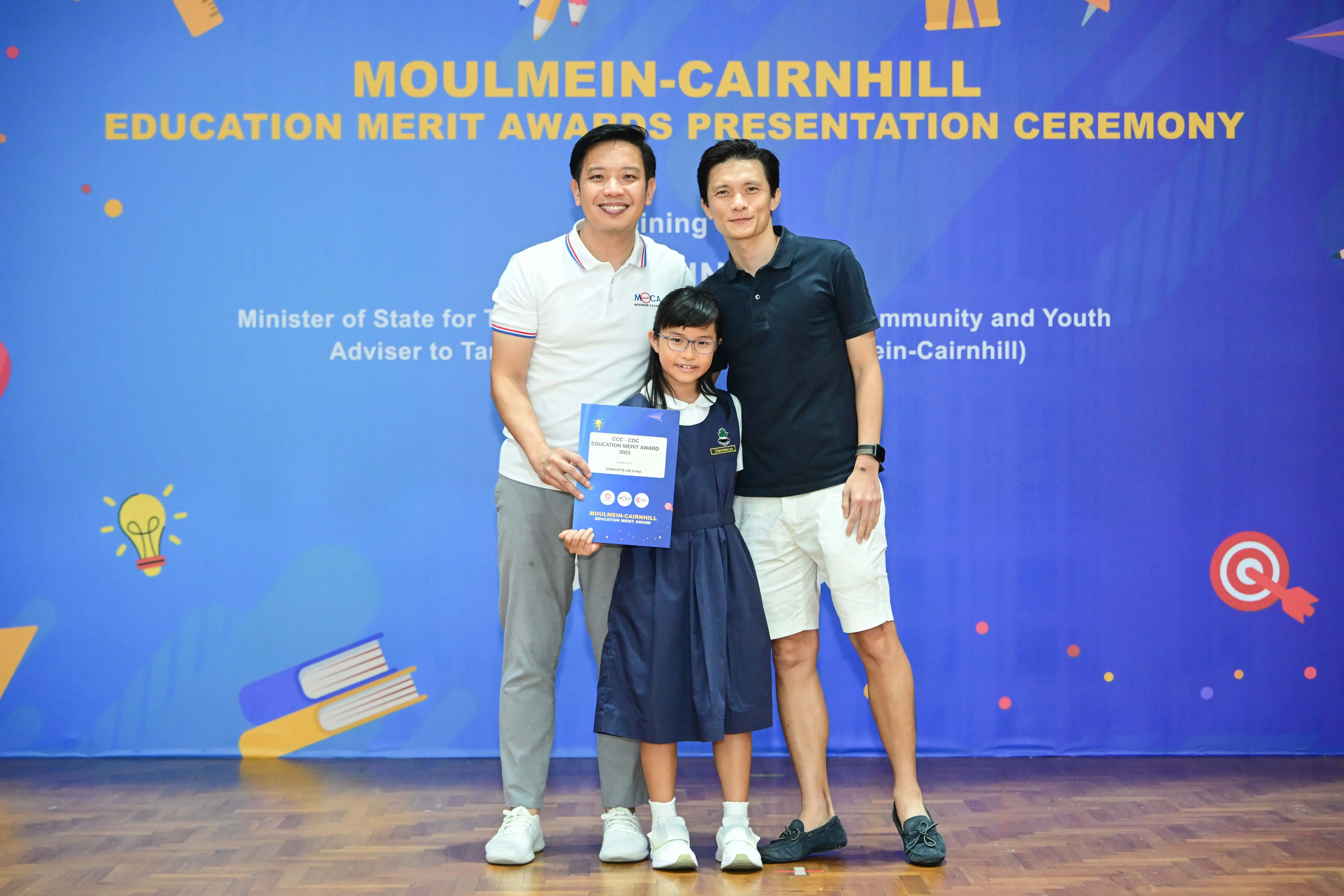 Two men and a young girl holding an award certificate pose before a Moulmein-Cairnhill Education Merit Awards banner.