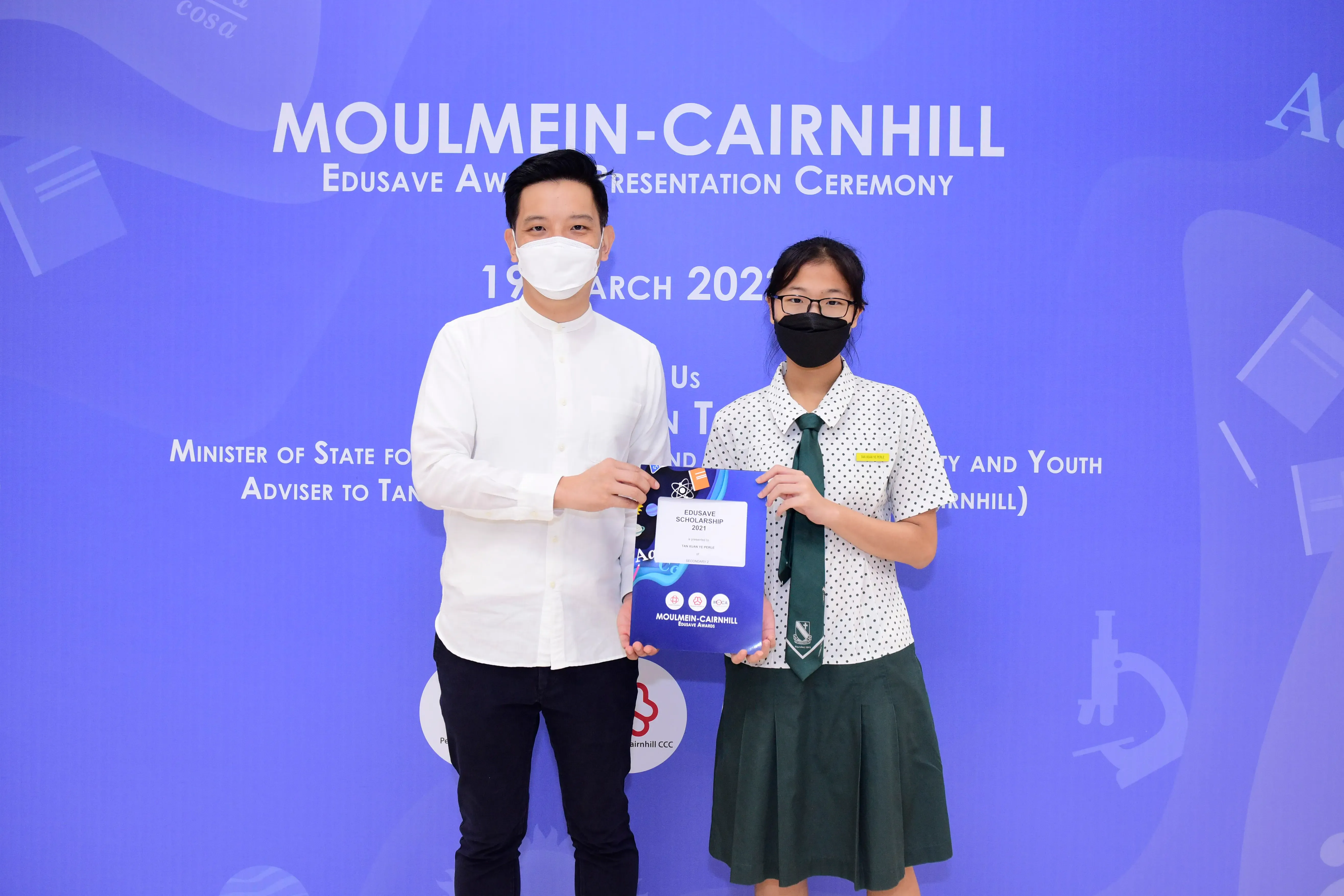 Two masked people hold an Edusave Scholarship award at the Moulmein-Cairnhill Edusave Awards Ceremony, March 2021.