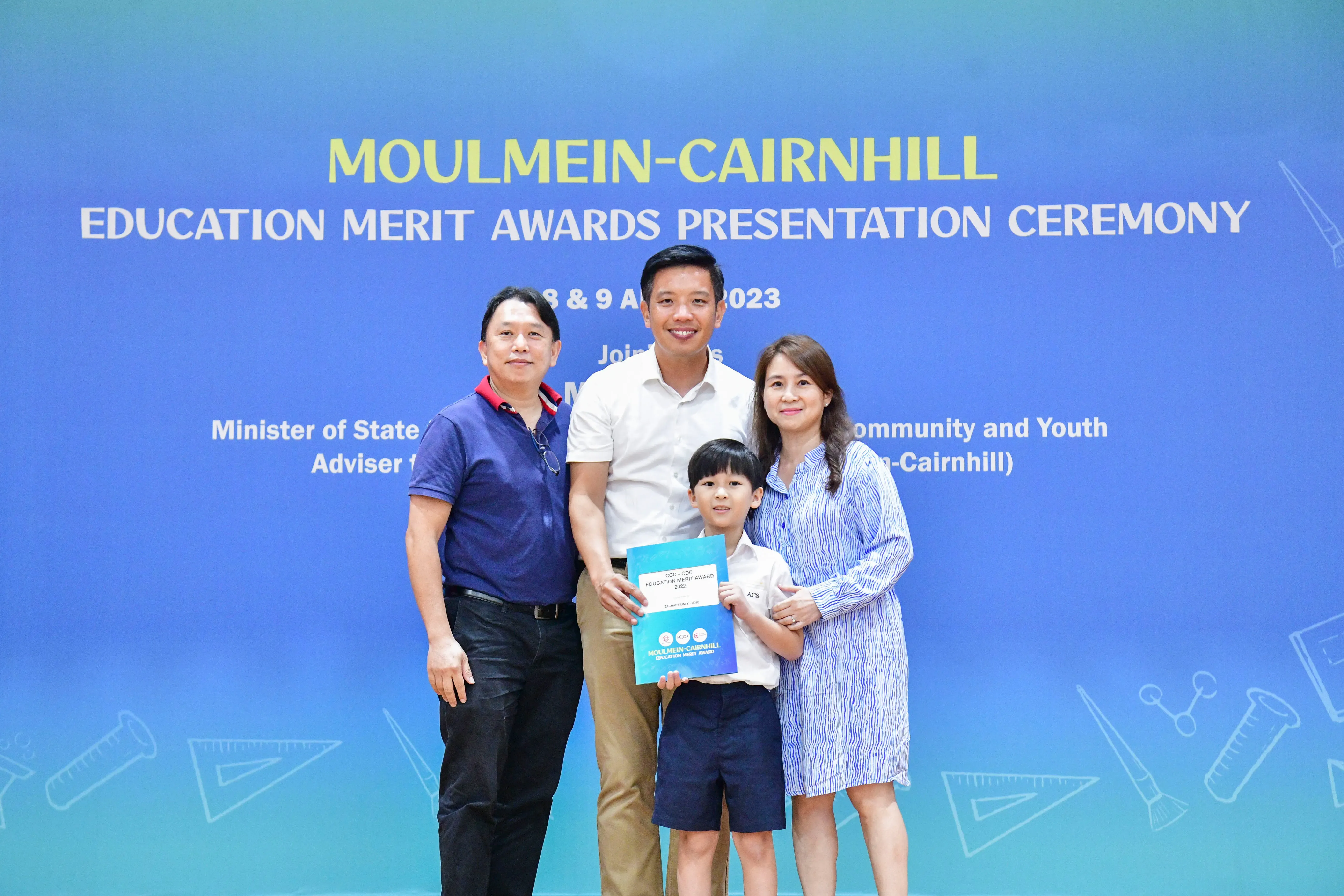 Three adults and a boy holding an award certificate pose before the Moulmein-Cairnhill Education Merit Awards banner.