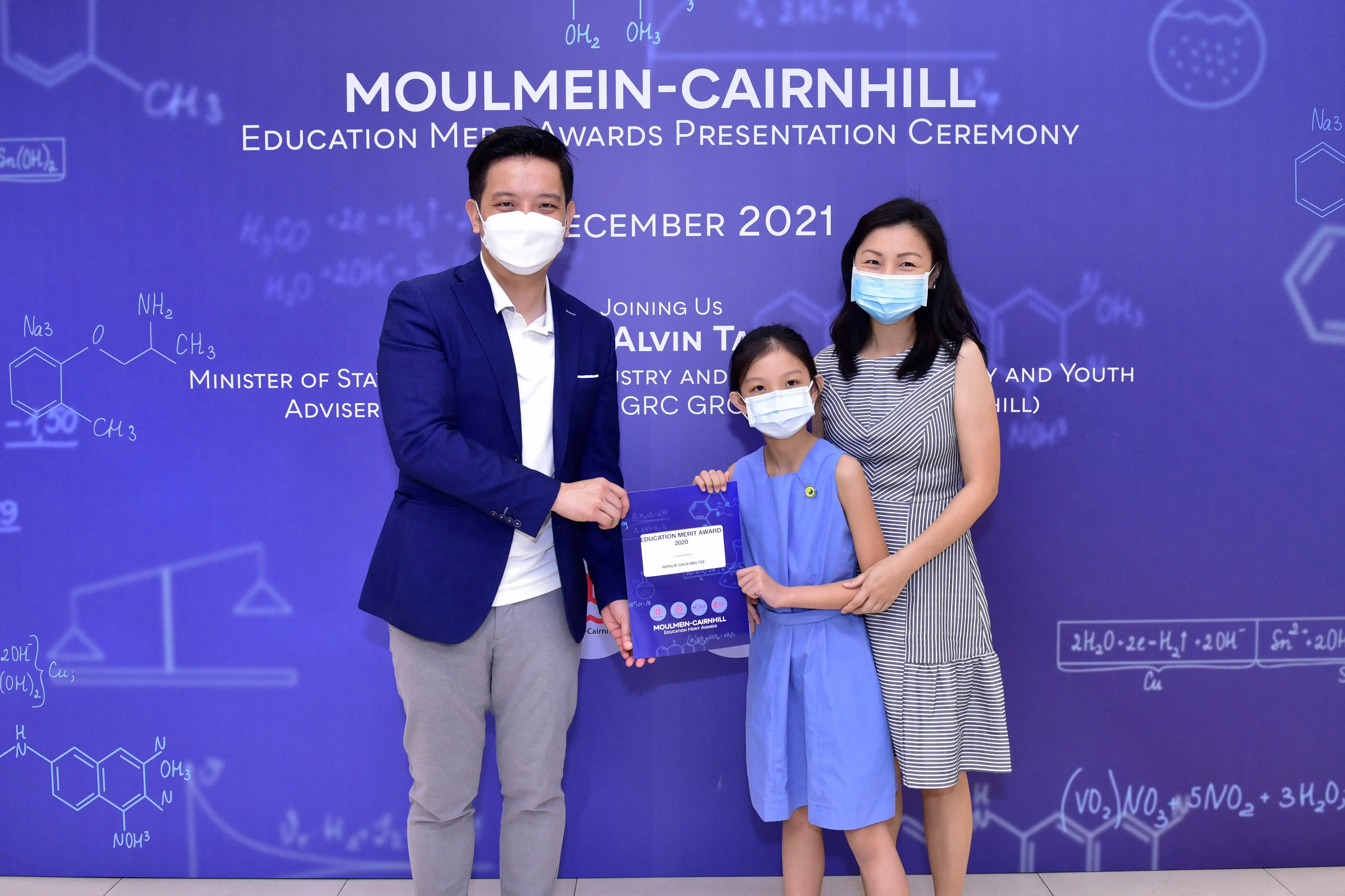 Two masked adults and a masked girl hold an award certificate at the Moulmein-Cairnhill Education Merit Awards 2021.