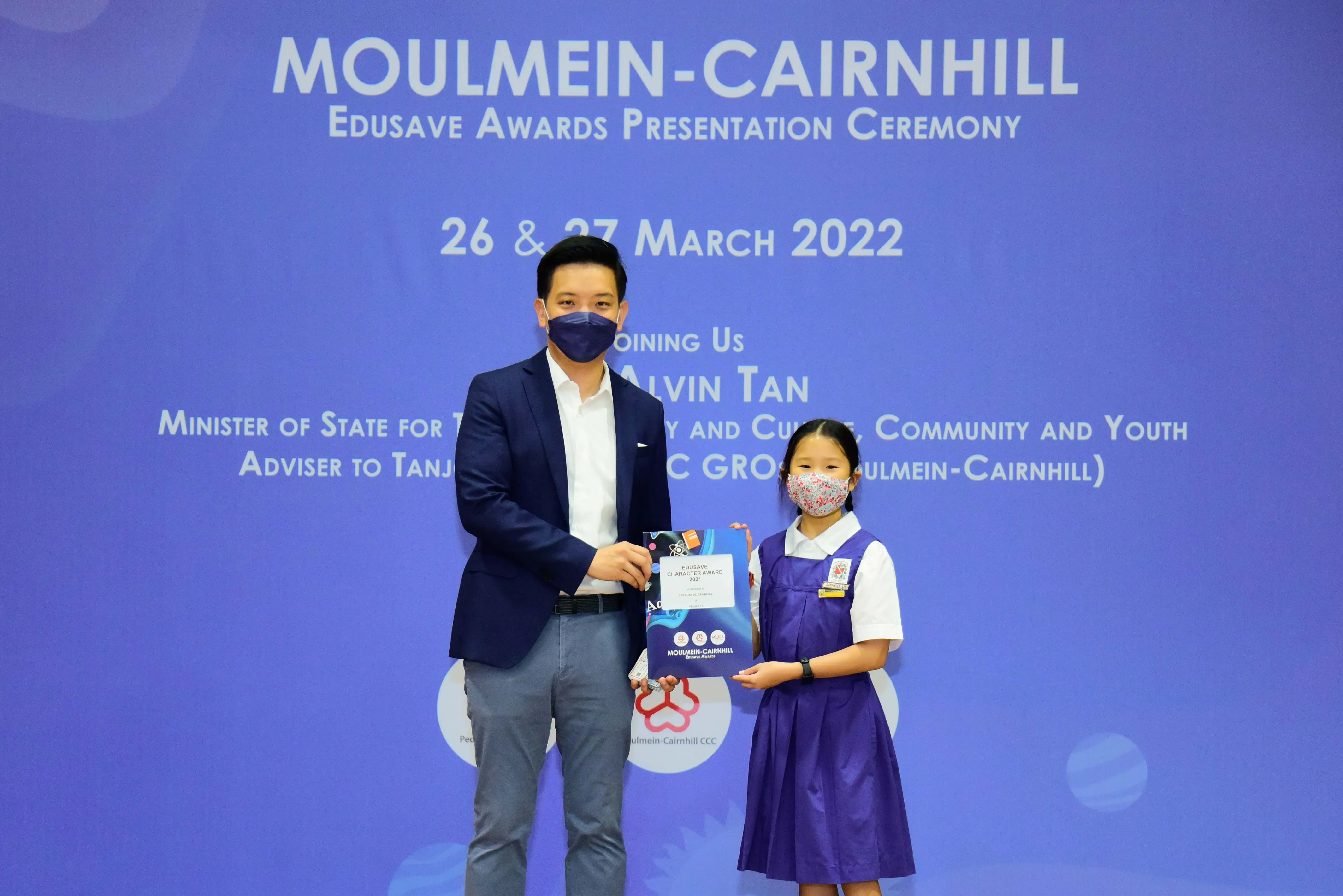 A man and a young girl in school uniform hold an Edusave award at the Moulmein-Cairnhill ceremony, March 2022.