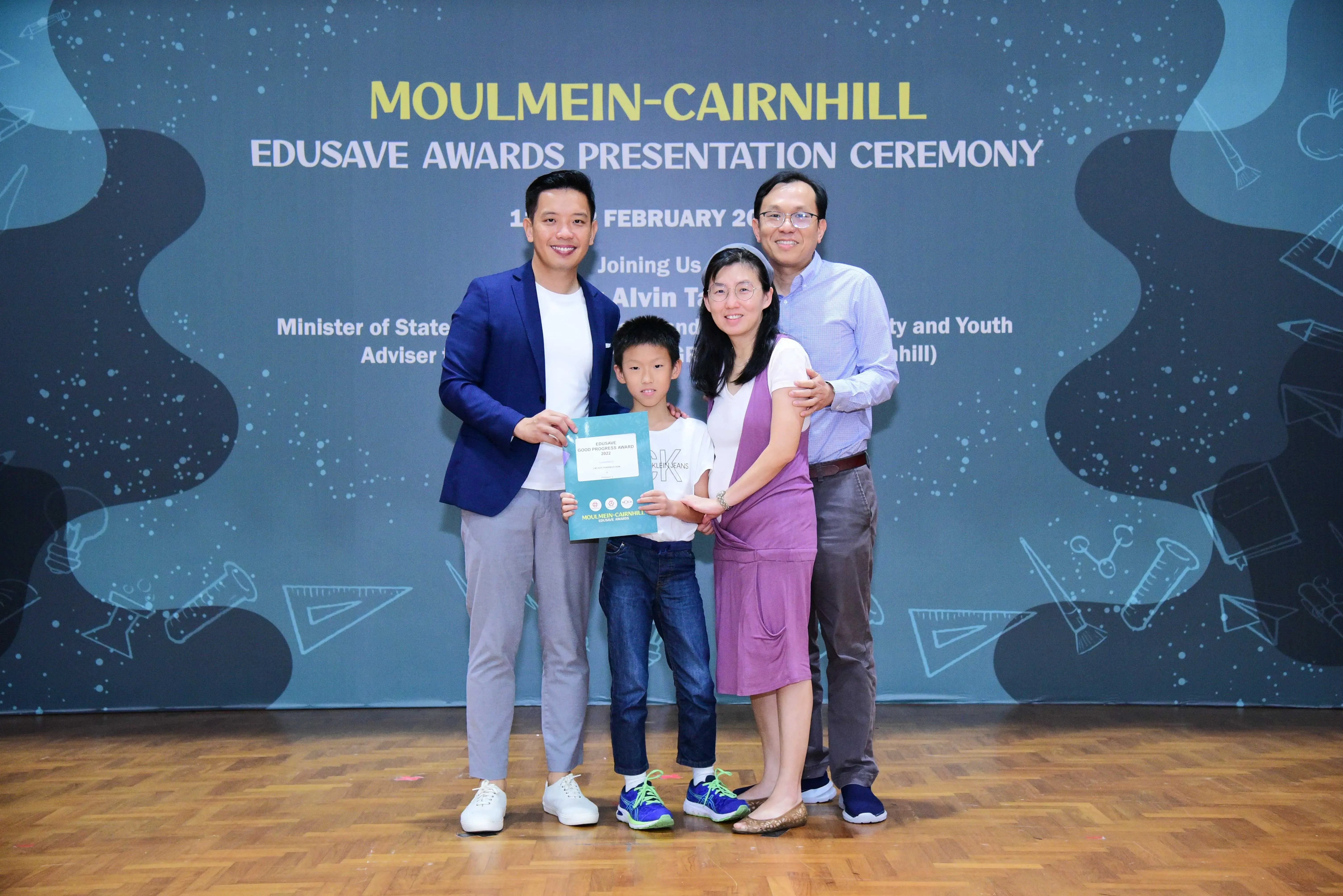 Four people pose at the Moulmein-Cairnhill Edusave Awards Presentation Ceremony, with a boy holding a certificate.