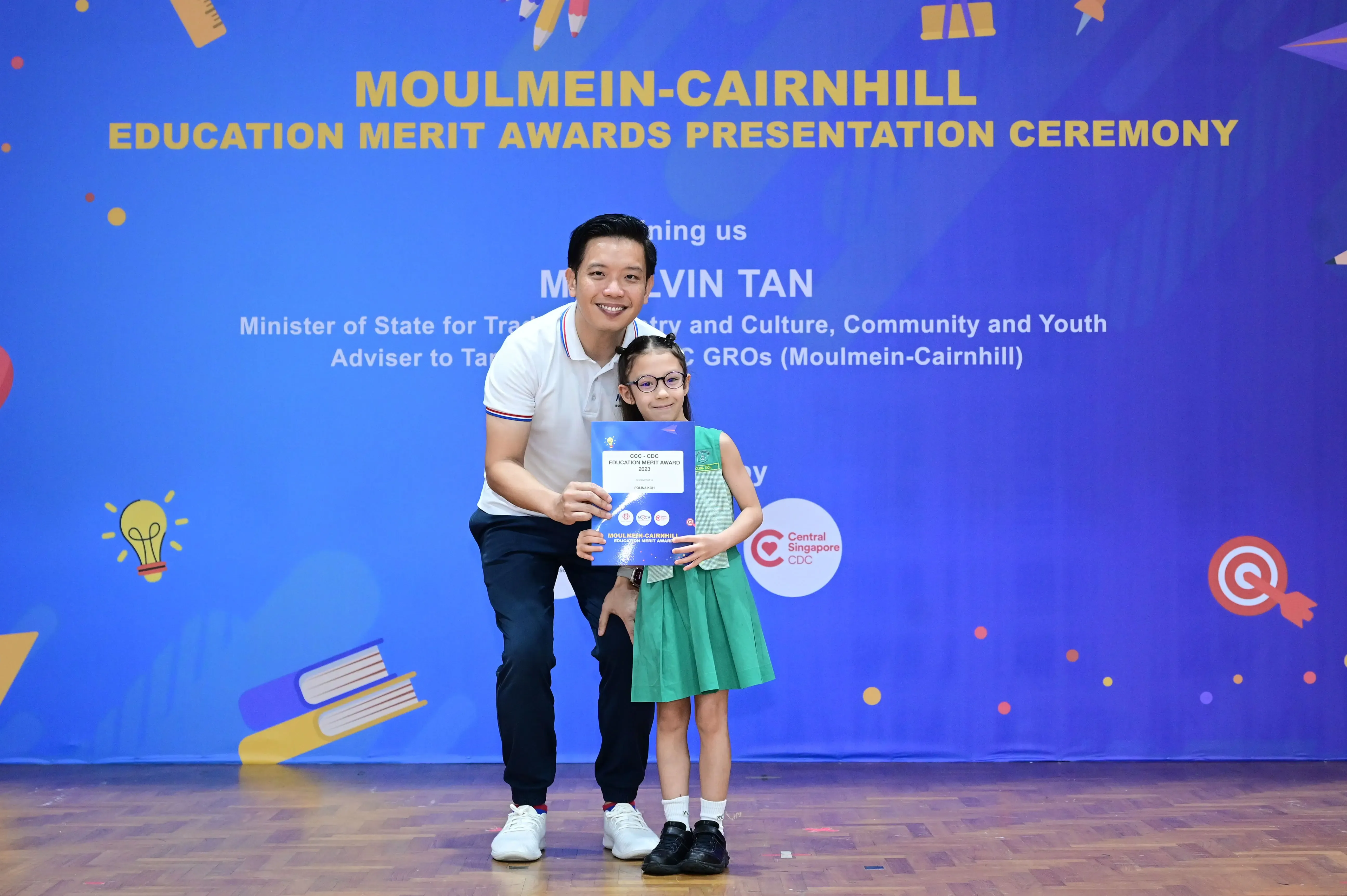 A man and a young girl holding an award certificate in front of a Moulmein-Cairnhill Education Merit Awards banner.