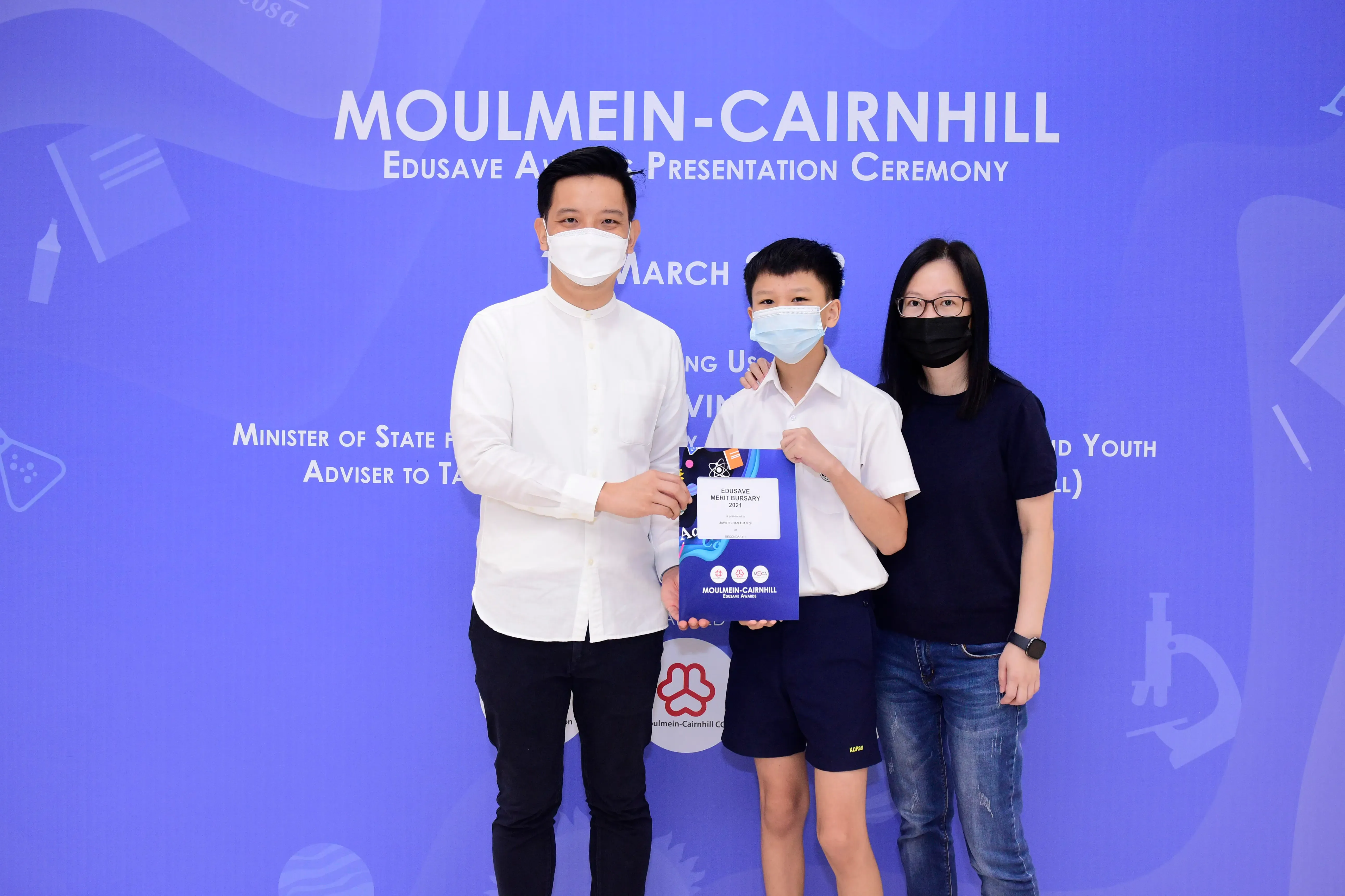 Three masked people pose at the Moulmein-Cairnhill Edusave Award Presentation Ceremony, a boy holds an award folder.