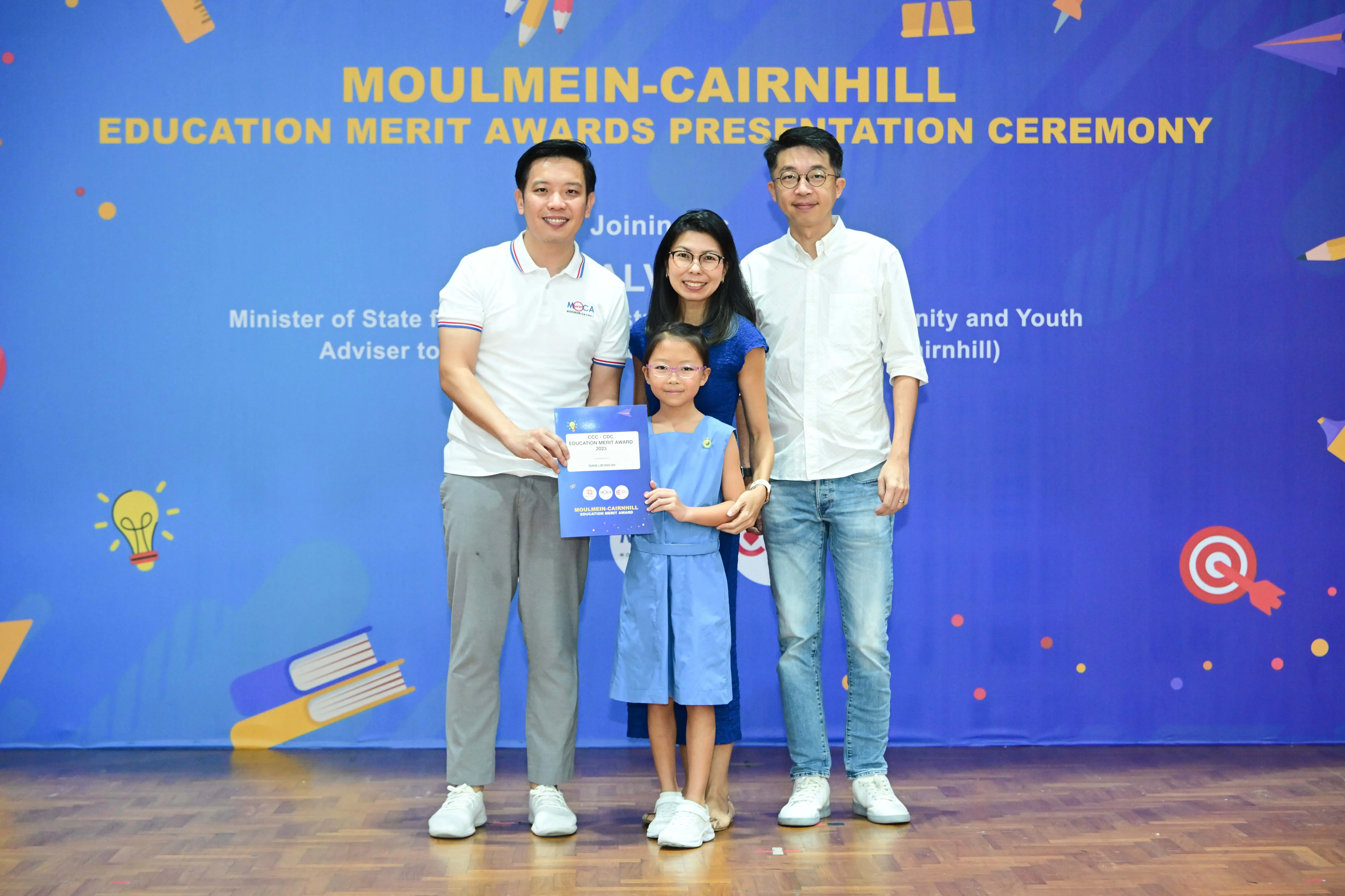 Three adults and a young girl holding a certificate pose before a Moulmein-Cairnhill Education Merit Awards banner.