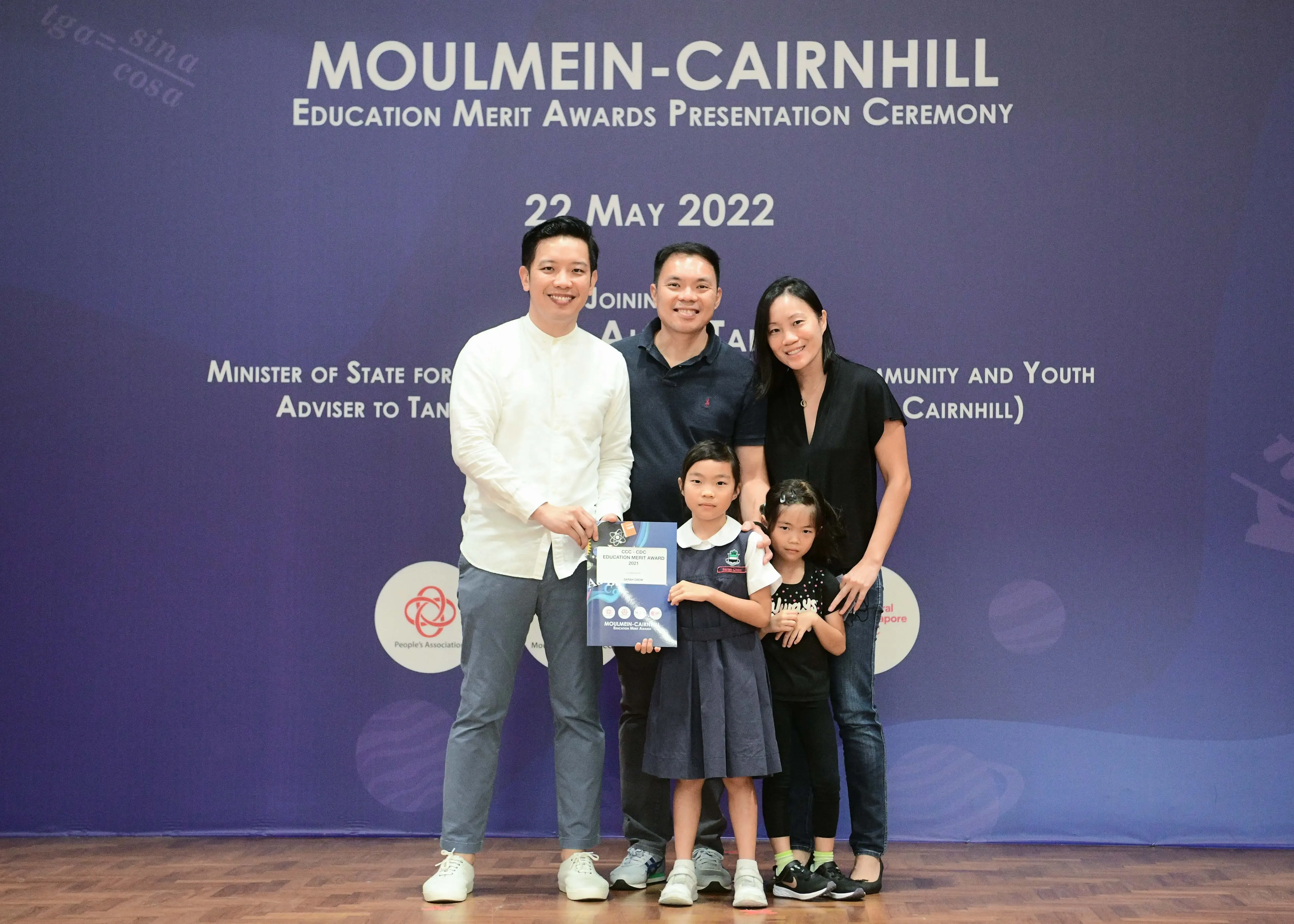 Five people pose before a Moulmein-Cairnhill Education Merit Awards banner dated 22 May 2022, a girl holding a certificate.