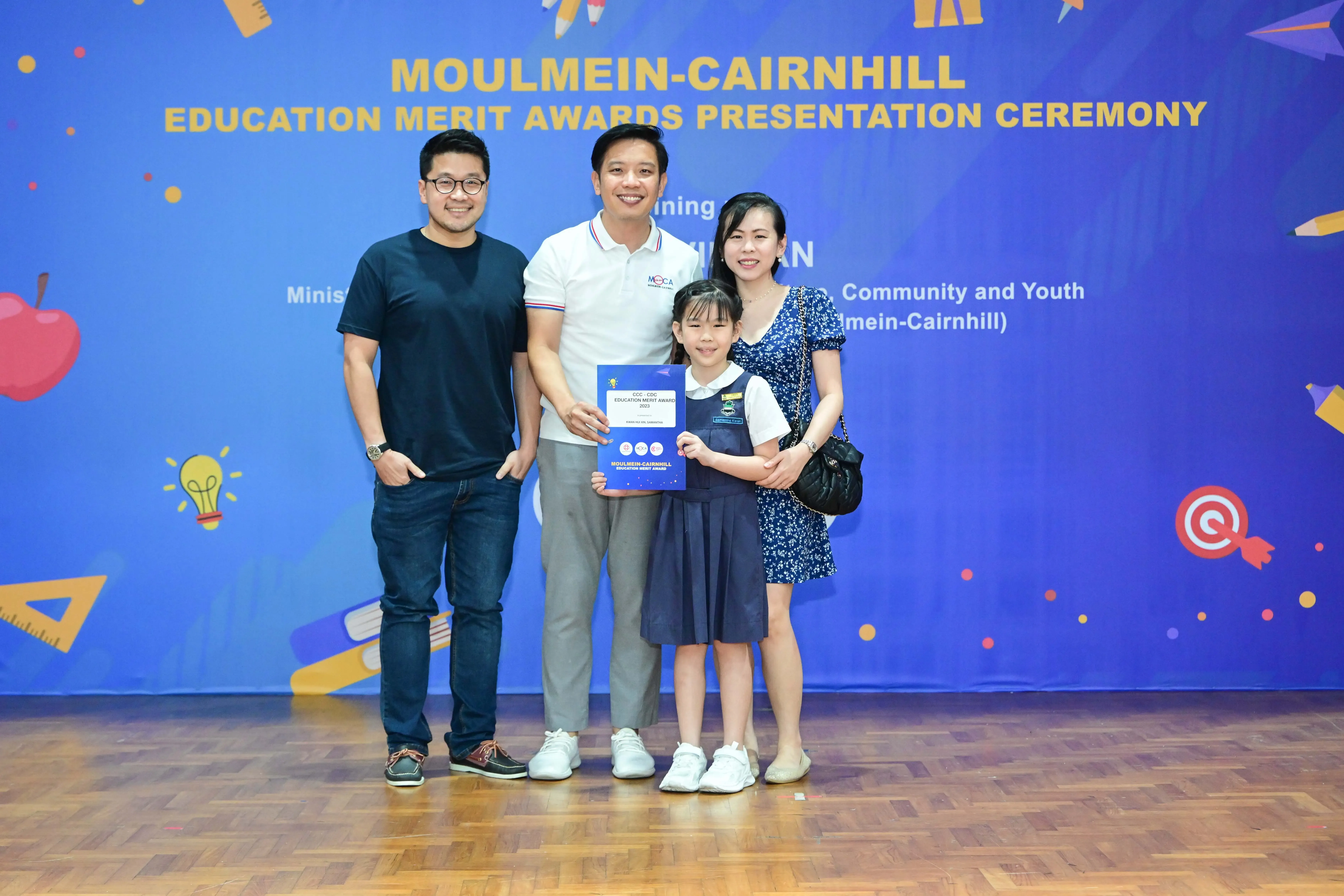 Three adults and a young girl hold an award certificate at the Moulmein-Cairnhill Education Merit Awards ceremony.