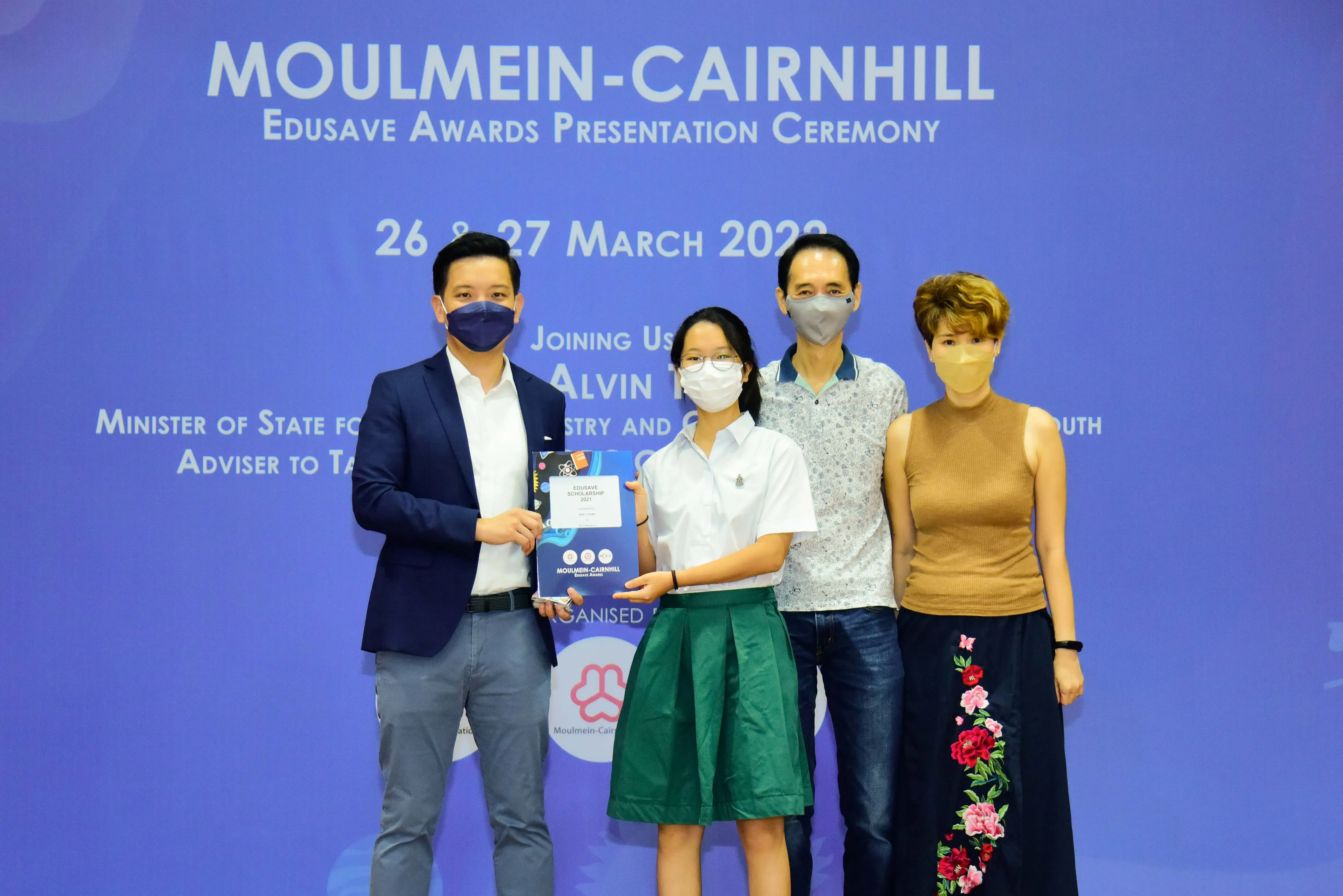 Four masked people pose at the Moulmein-Cairnhill Edusave Awards Presentation Ceremony, with a girl holding an award.