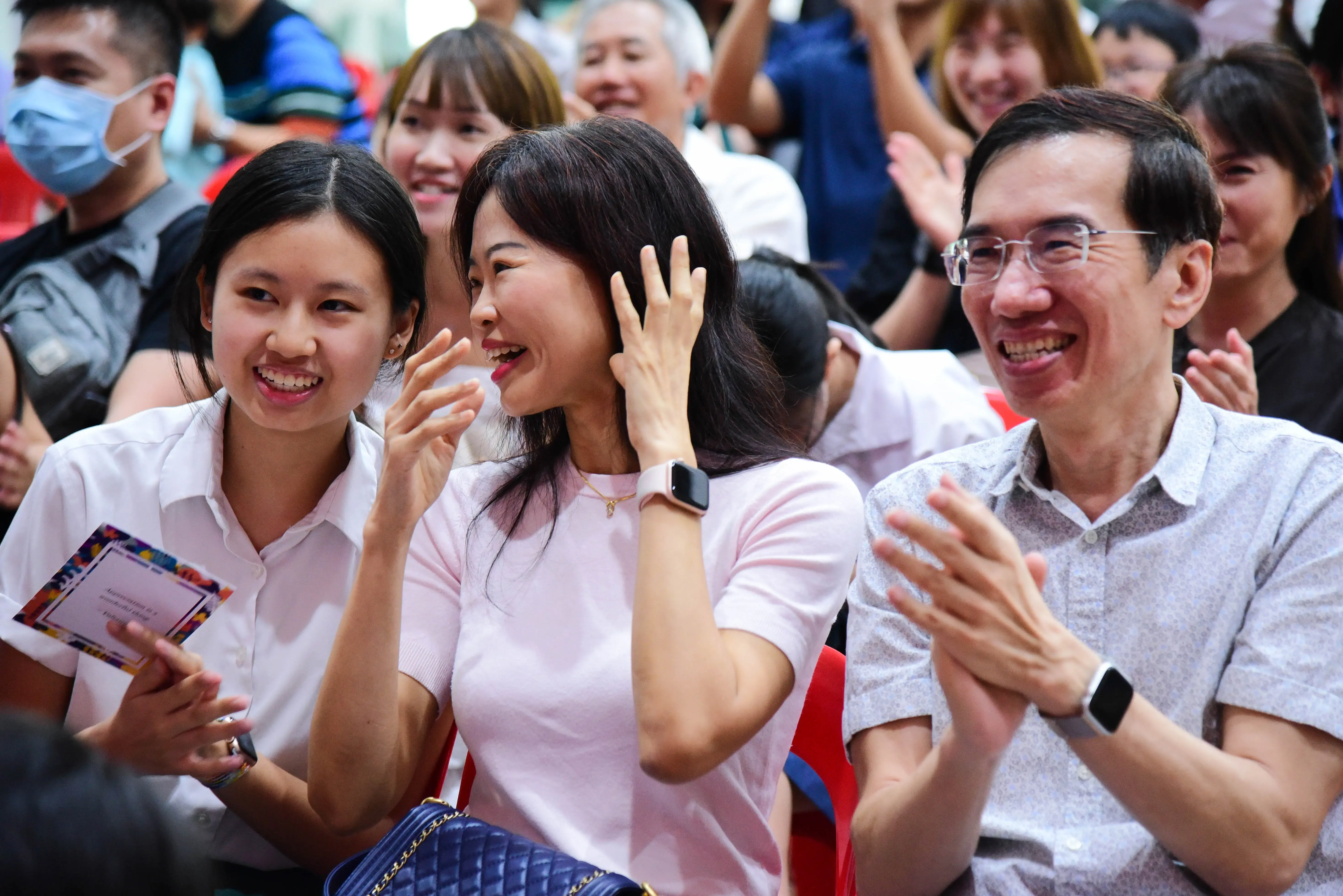 Three smiling Asian people clapping in a crowd, a teenage girl, a woman in pink, and a man with glasses.