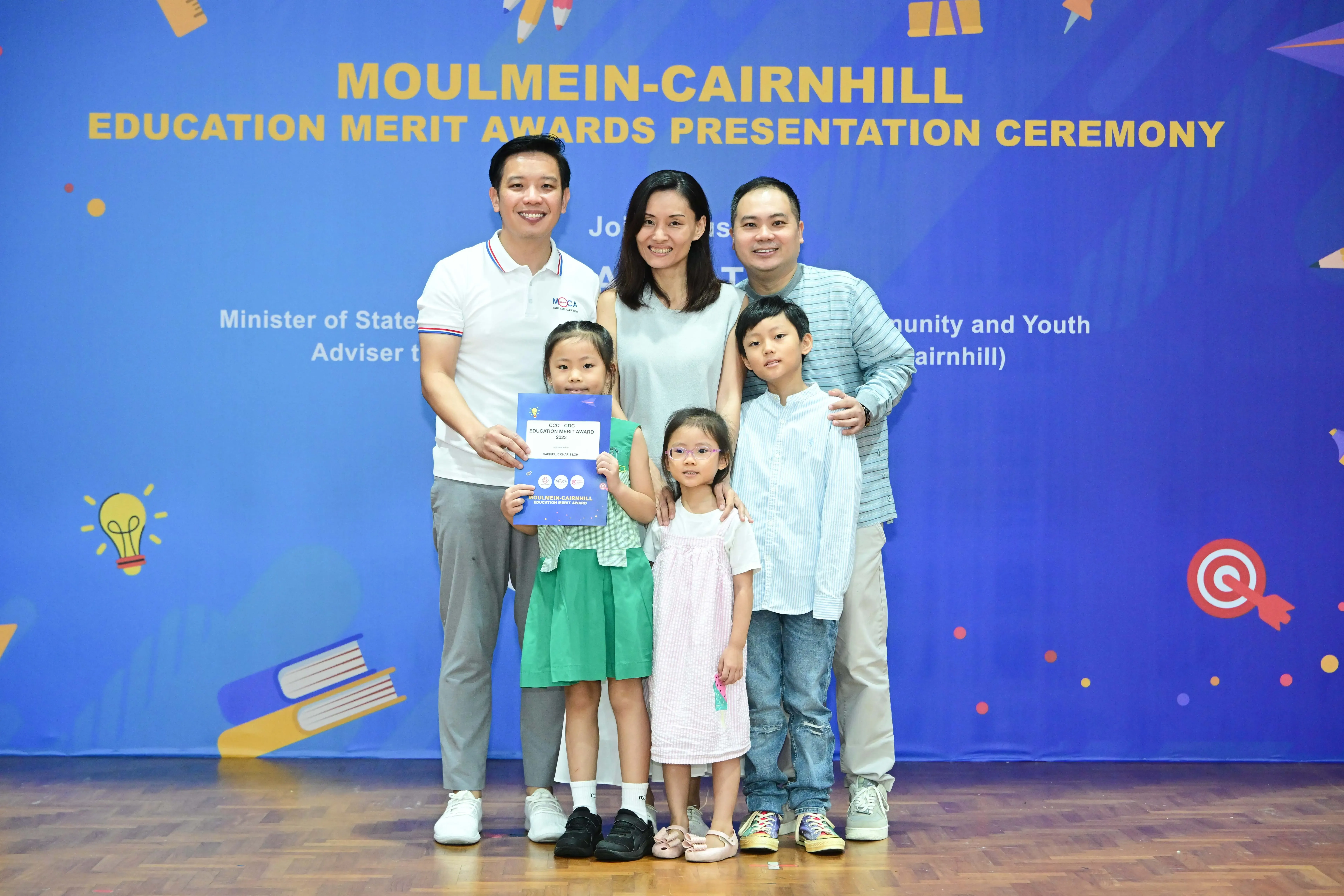 Family of five posing in front of a blue Moulmein-Cairnhill Education Merit Awards banner, child holding a certificate.