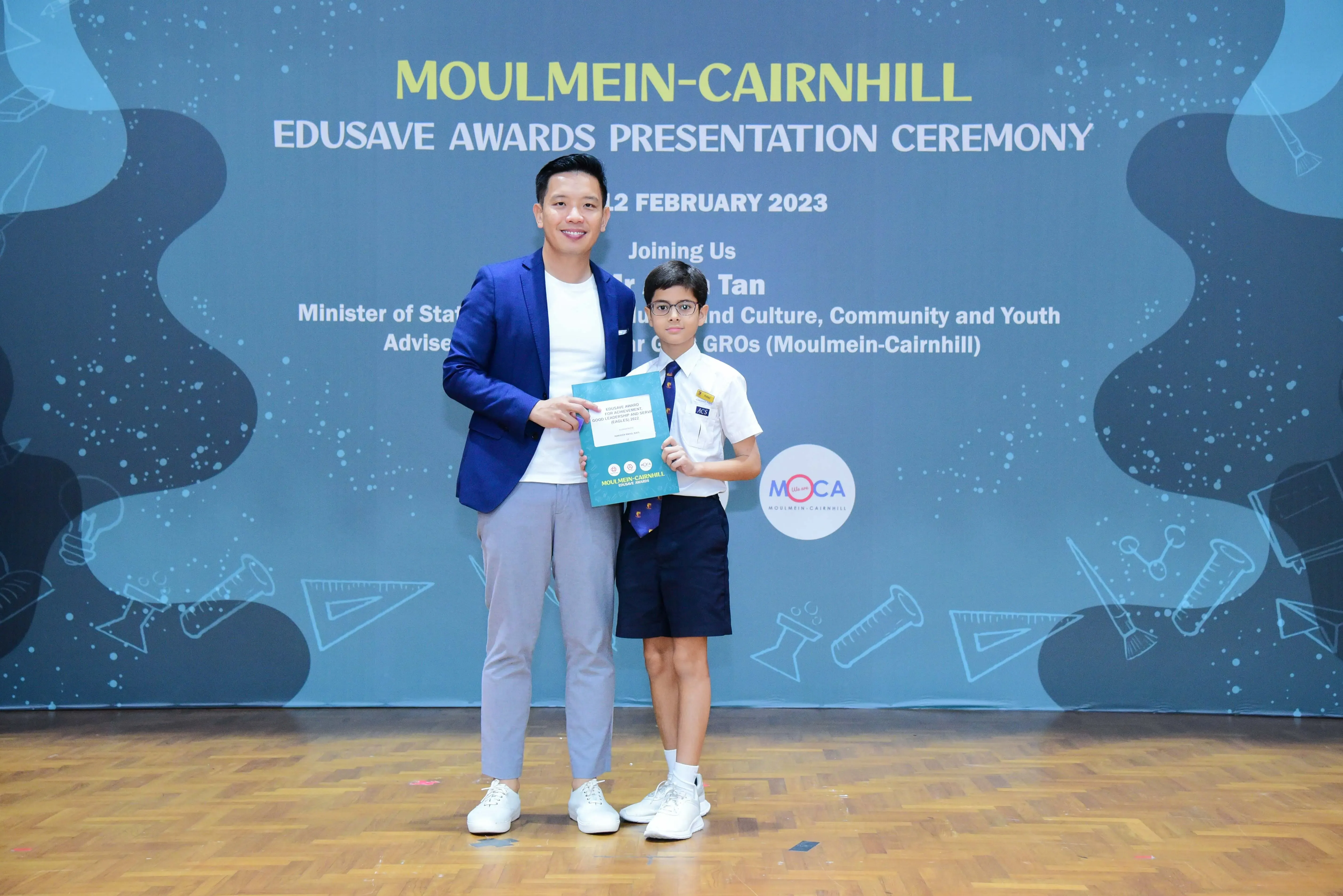 An adult in a blue blazer and a young student in uniform hold a certificate at the Moulmein-Cairnhill Edusave Awards 2023.