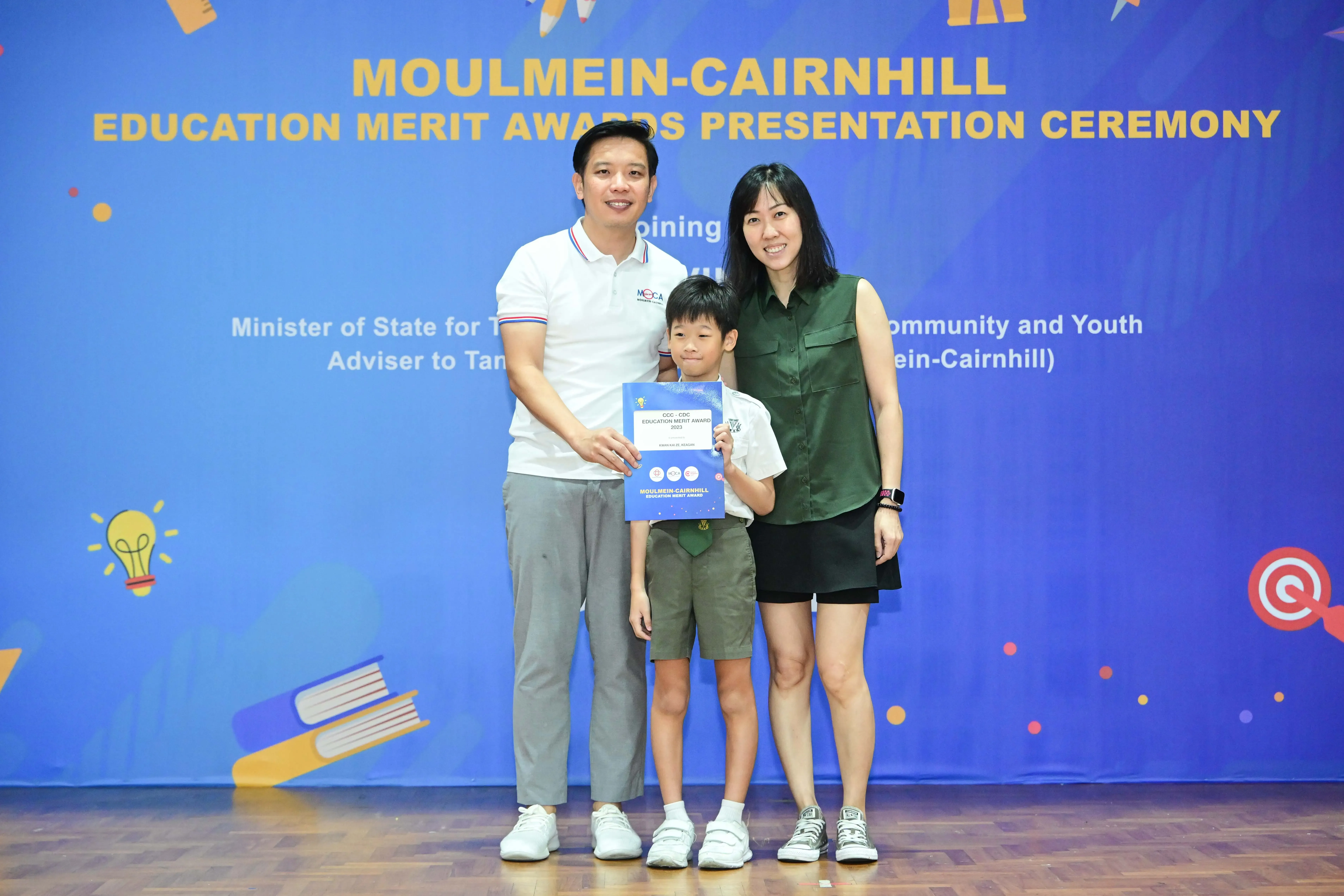 A boy holding an award certificate stands between two adults at the Moulmein-Cairnhill Education Merit Awards Ceremony.