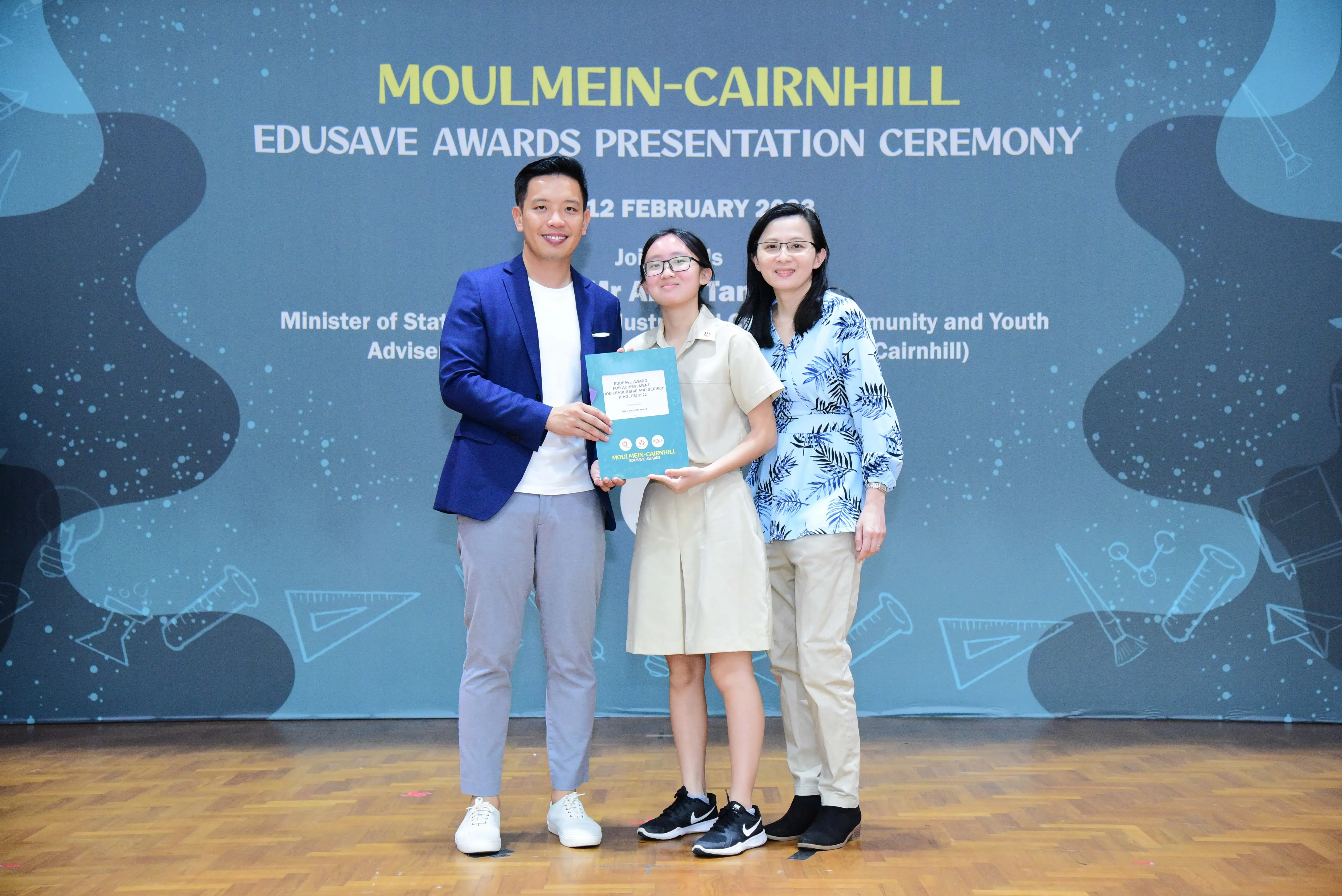 Three people smile while holding an award booklet at the Moulmein-Cairnhill Edusave Awards Presentation Ceremony.