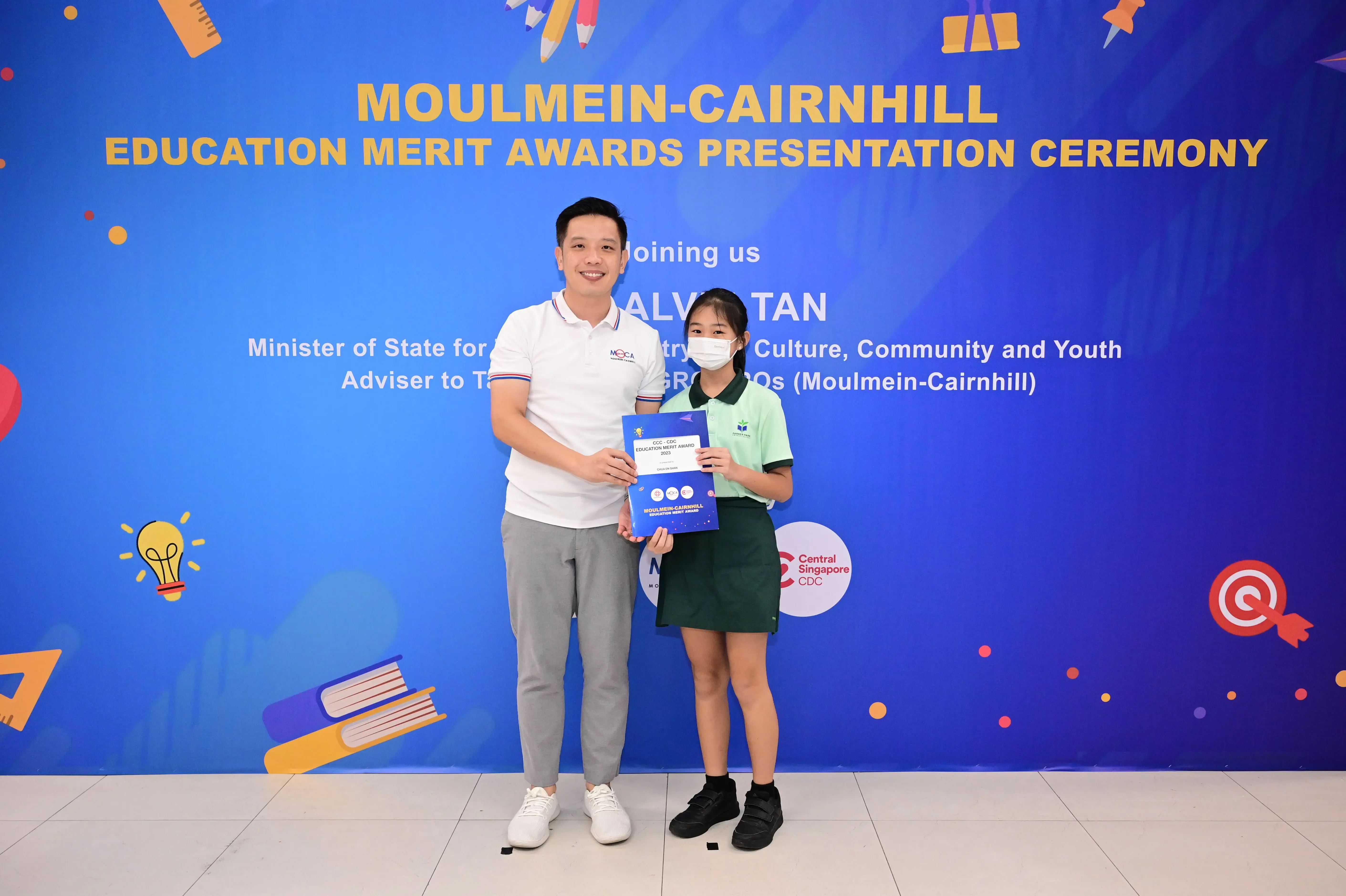 A man and a young girl hold an award certificate in front of a Moulmein-Cairnhill Education Merit Awards banner.