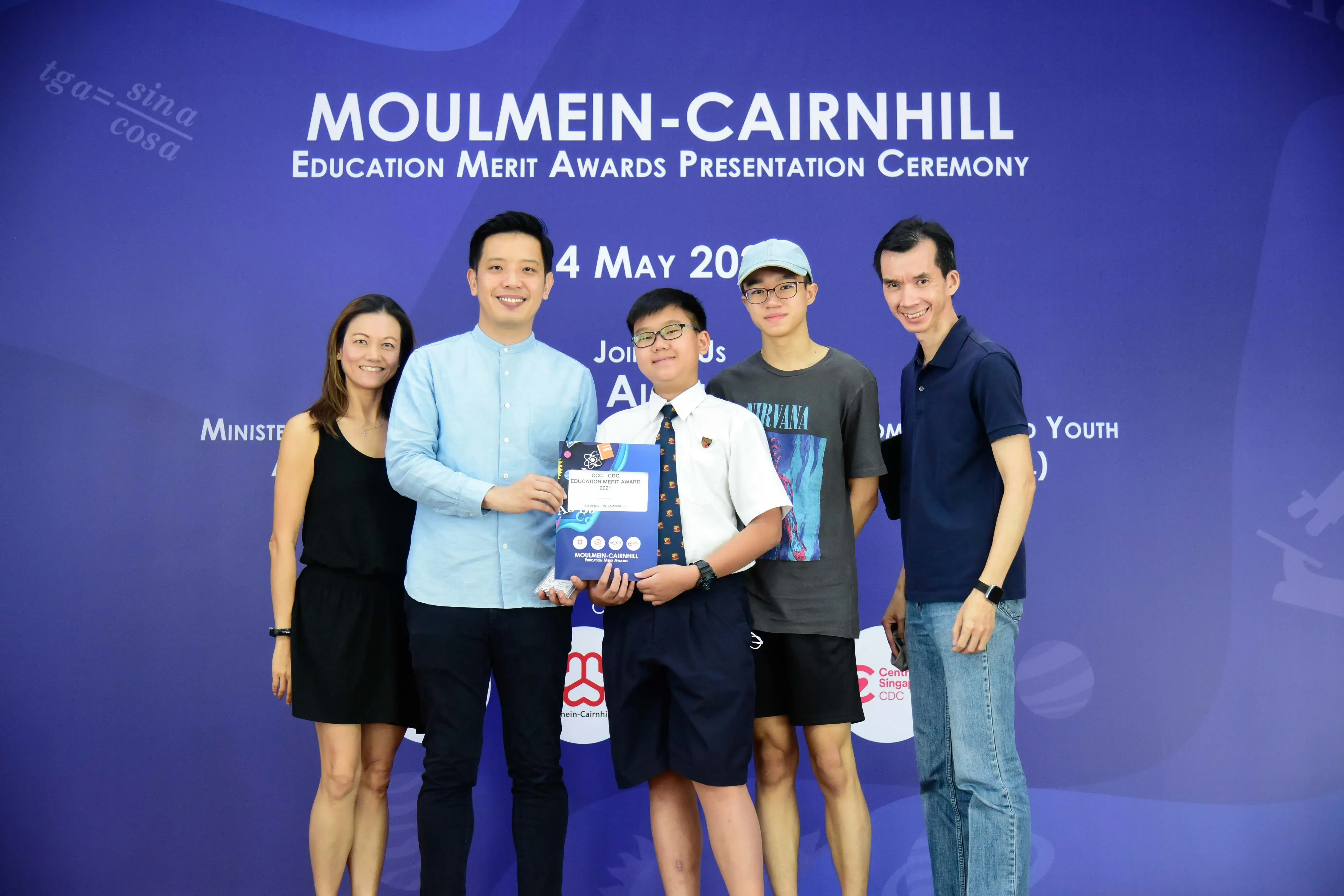 Five people pose together at the Moulmein-Cairnhill Education Merit Awards Presentation Ceremony, with a boy holding an award folder.