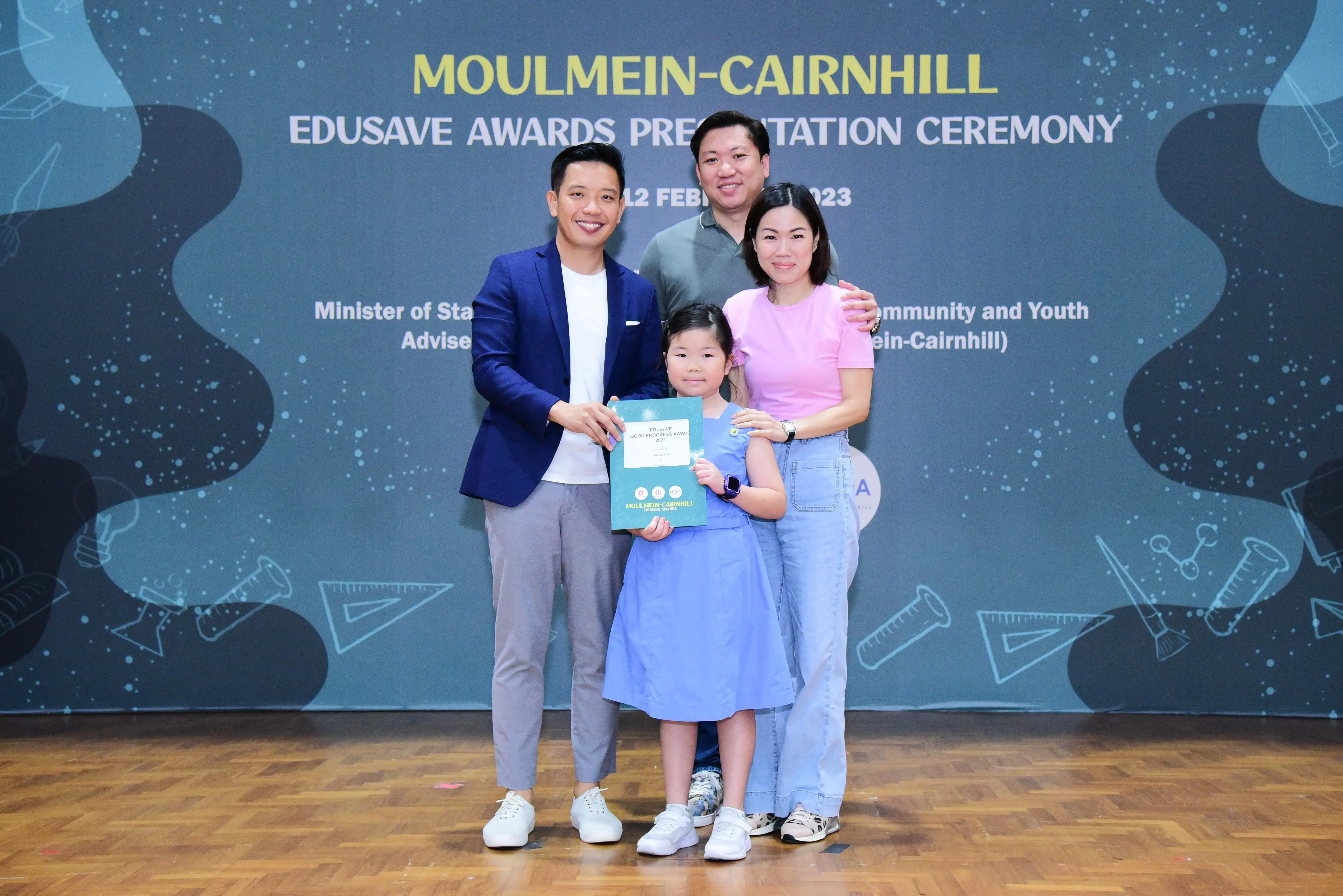 Four people pose at the Moulmein-Cairnhill Edusave Awards Ceremony, with a young girl holding a teal certificate.