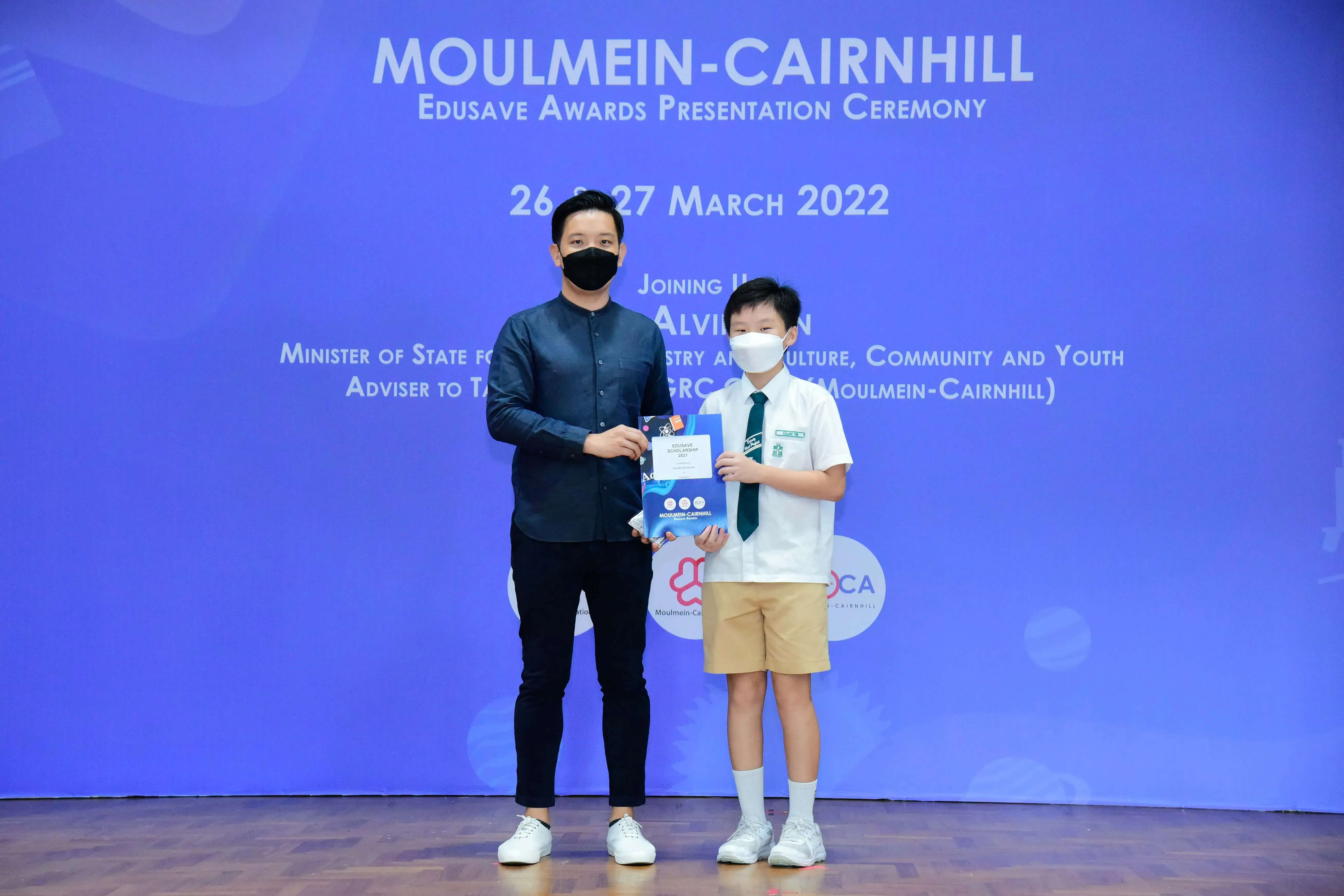 An adult and a young student in uniform hold an award folder at the Moulmein-Cairnhill Edusave ceremony, March 2022.